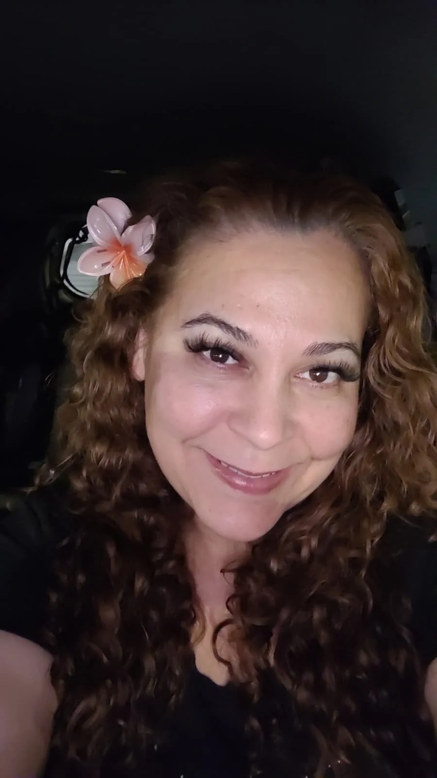 A smiling woman with curly brown hair and a pink flower in her hair.