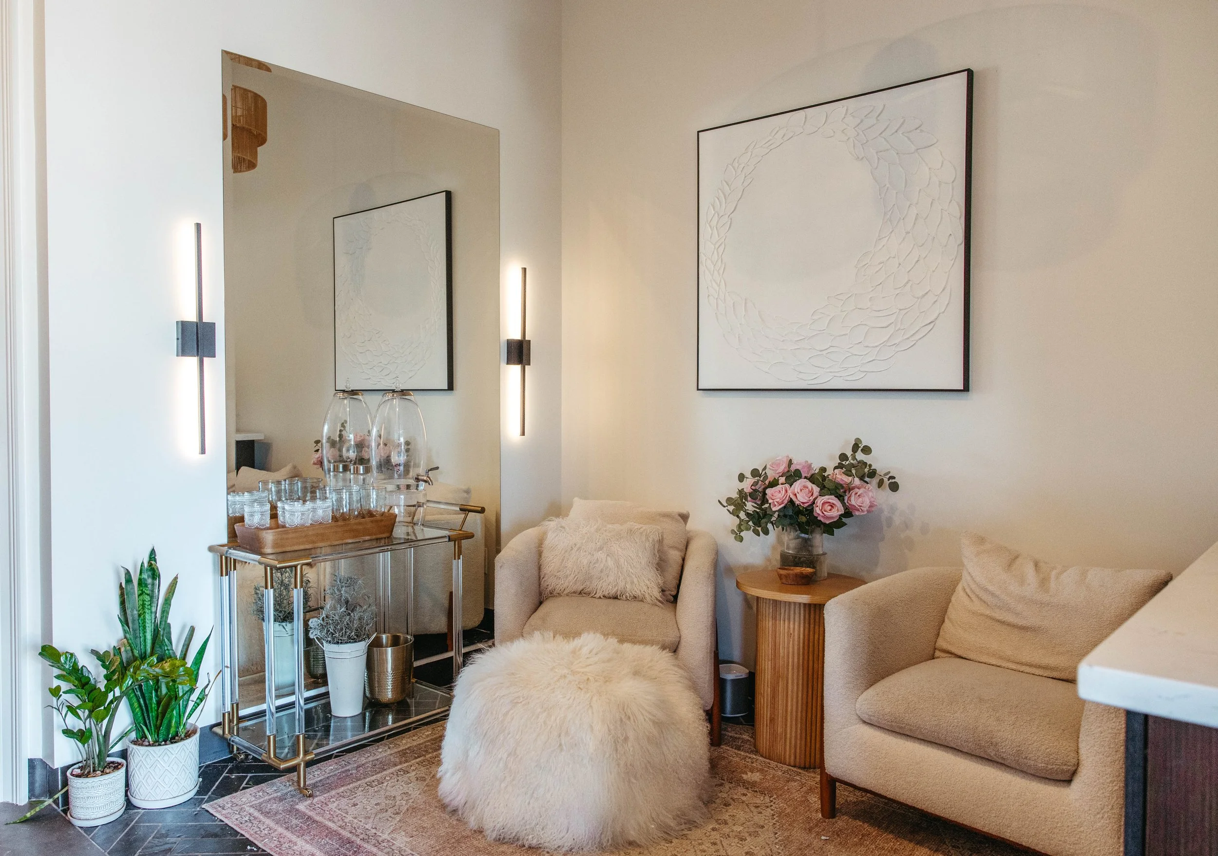 A cozy living room corner with beige armchairs, a fluffy ottoman, a round wooden side table with pink roses, a big framed abstract art piece, a metal and glass bar cart with glassware, a mirror on the wall, and decorative plants on the floor.