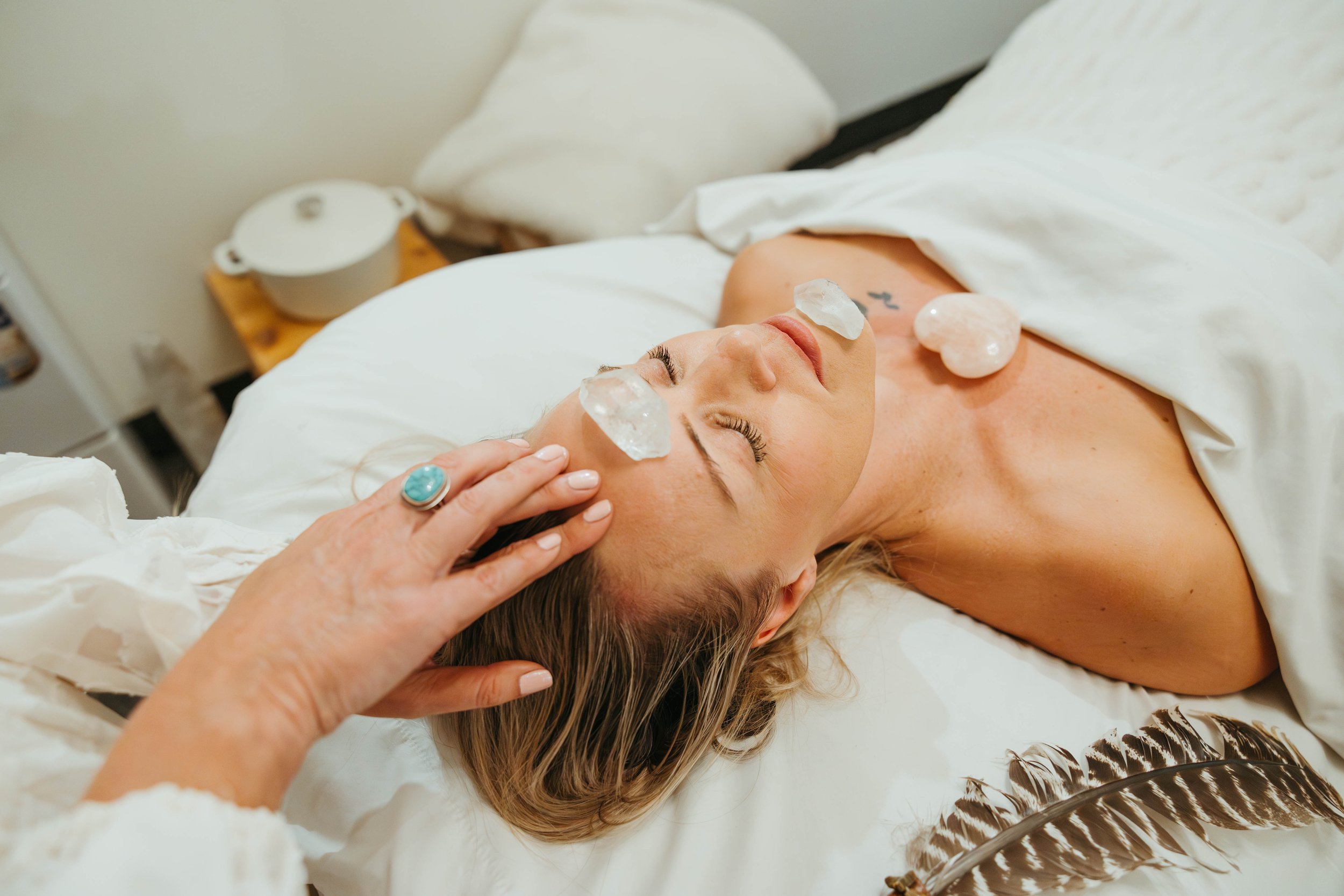 A woman is lying on a bed with her eyes closed, receiving a crystal facial treatment. She has crystals placed on her forehead and cheeks, and a person with a turquoise ring is gently touching her head.