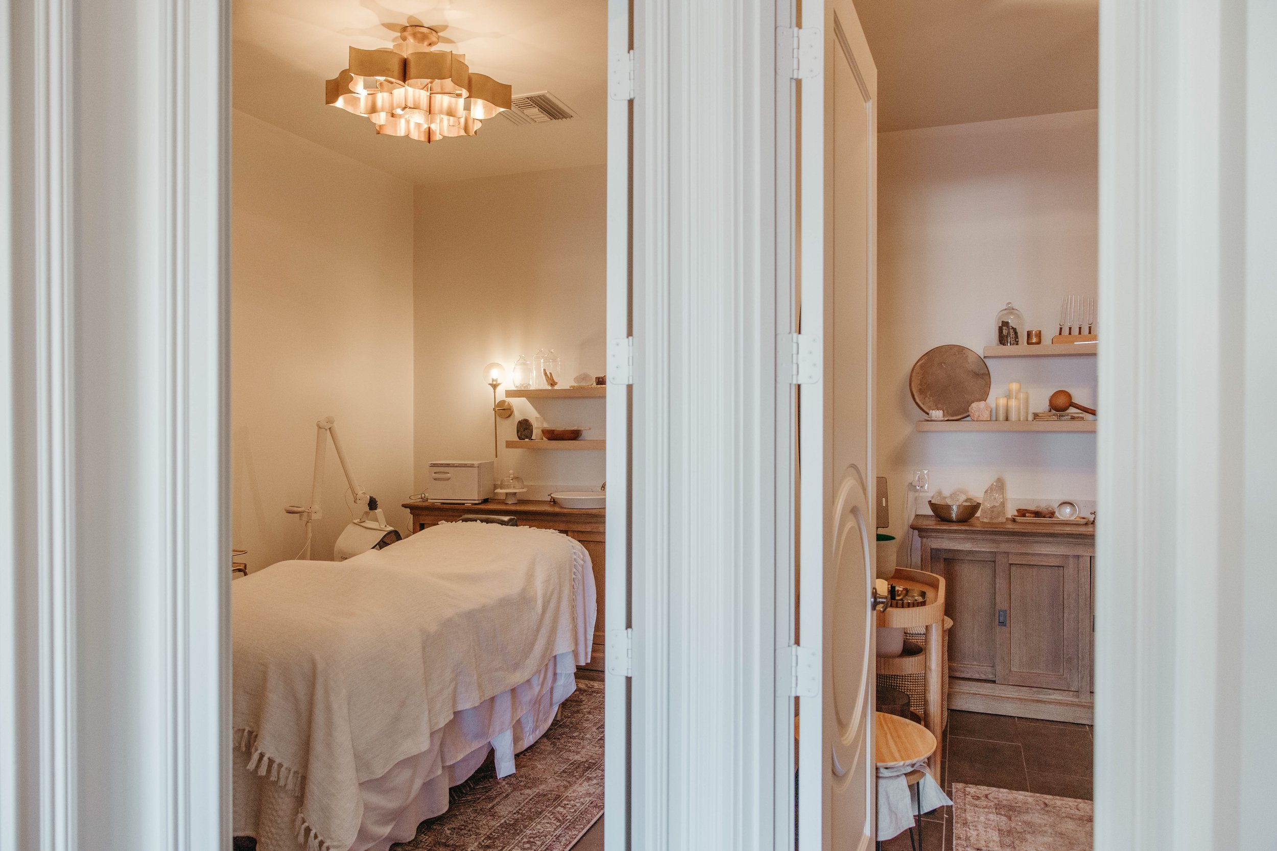 A view through a partially open white louvered door shows a cozy room decorated in neutral tones, with a massage or treatment bed, shelves with candles and decorative objects, and warm lighting from a pendant lamp and wall sconce.