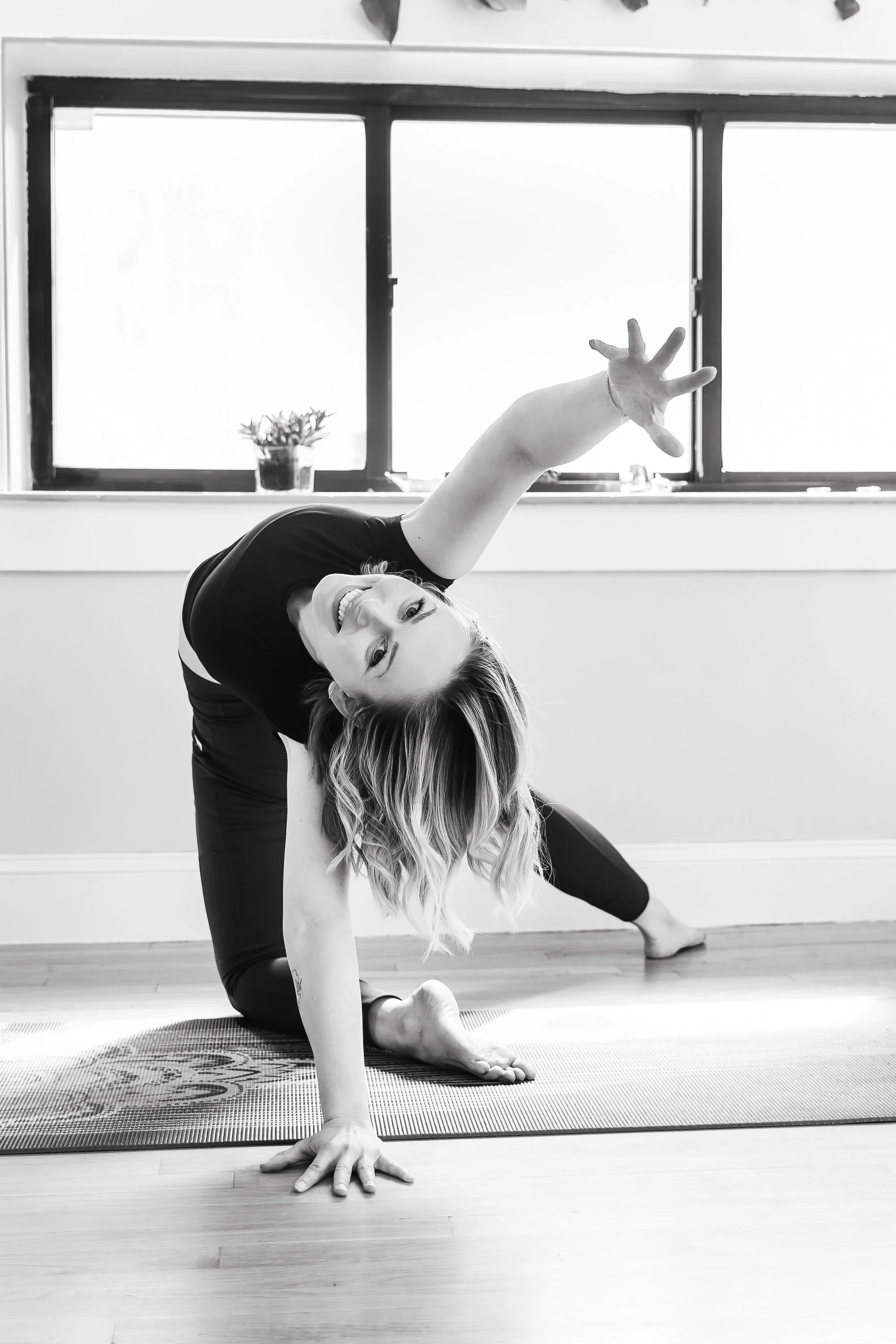 A woman practicing yoga indoors, performing a pose on a mat near a window, with a smiling expression.