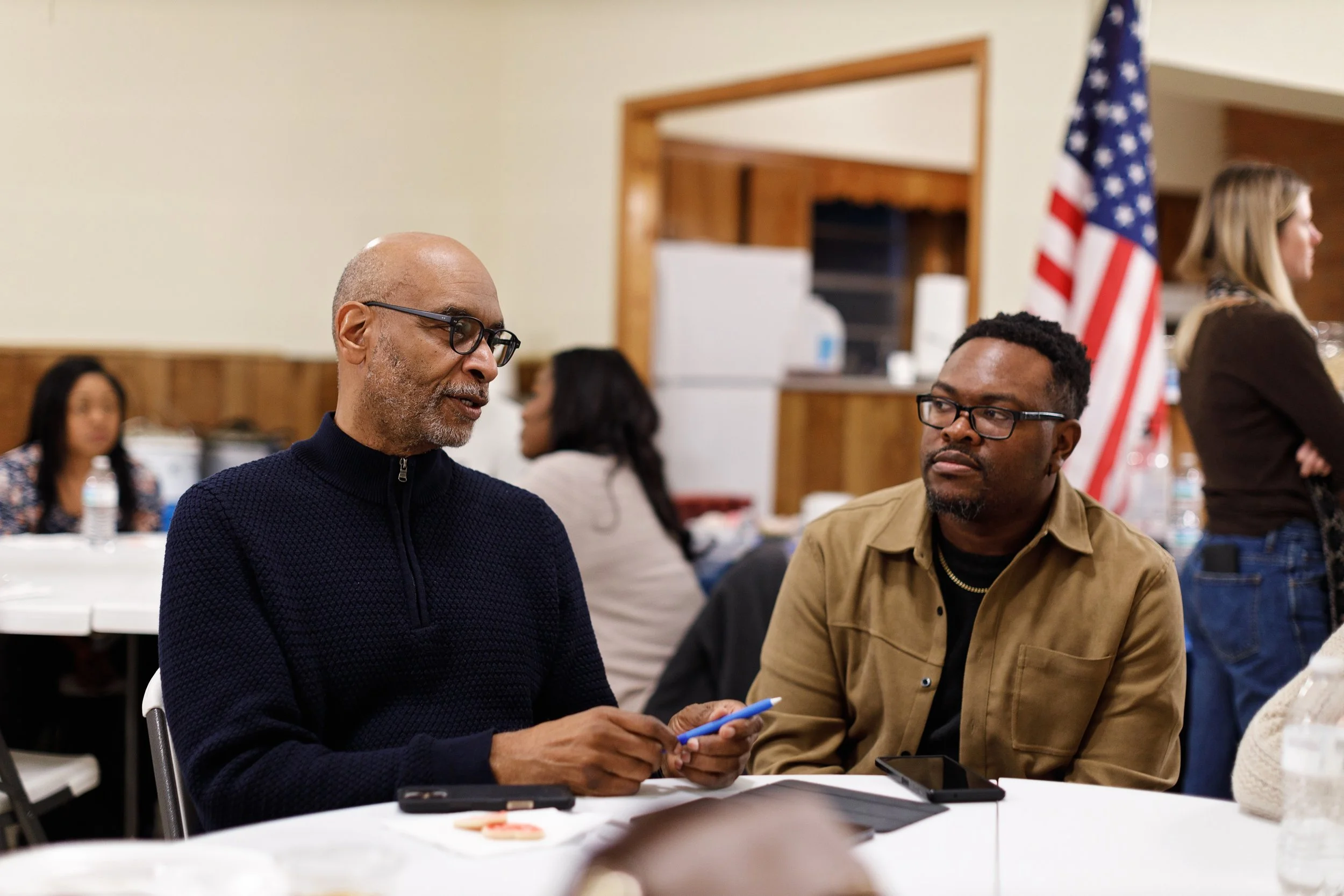 Two men engaged in conversation at a table during a gathering, with American flag in the background.