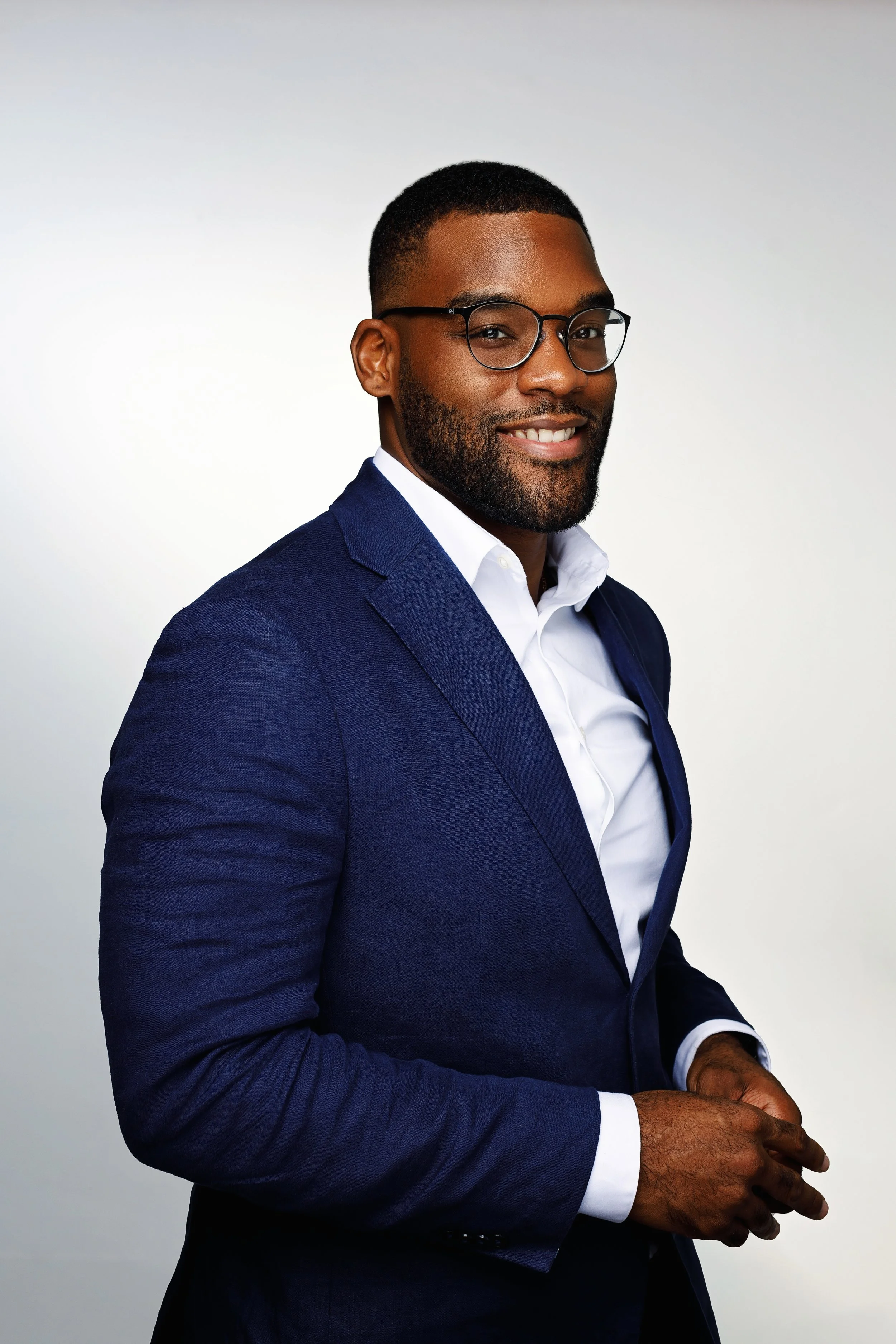 A smiling Black man wearing glasses, a white dress shirt, and a blue suit, standing against a white background.