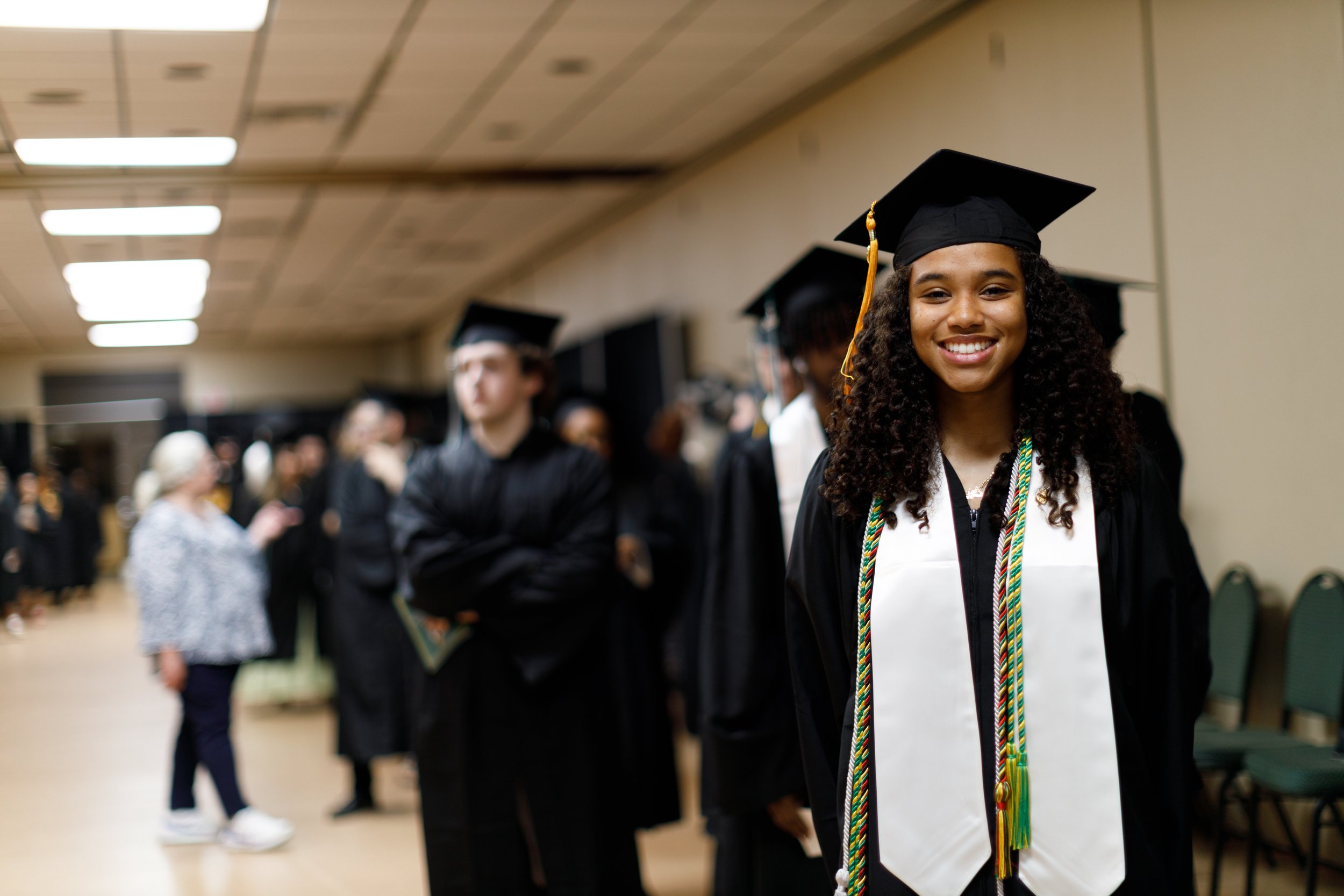 A young woman in graduation cap and gown smiling at a graduation ceremony with other graduates in the background.