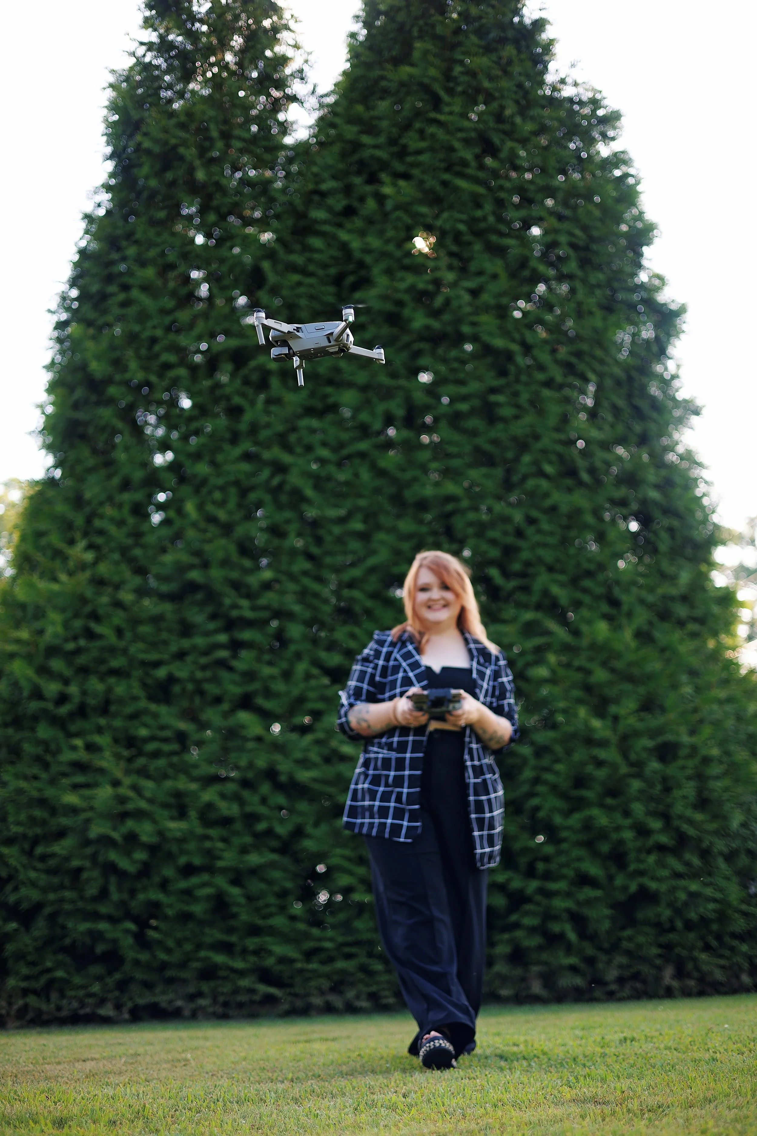 A woman flying a drone outdoors near a tall, leafy tree. She is holding a remote control, smiling, and wearing a dark checkered blazer and black pants.