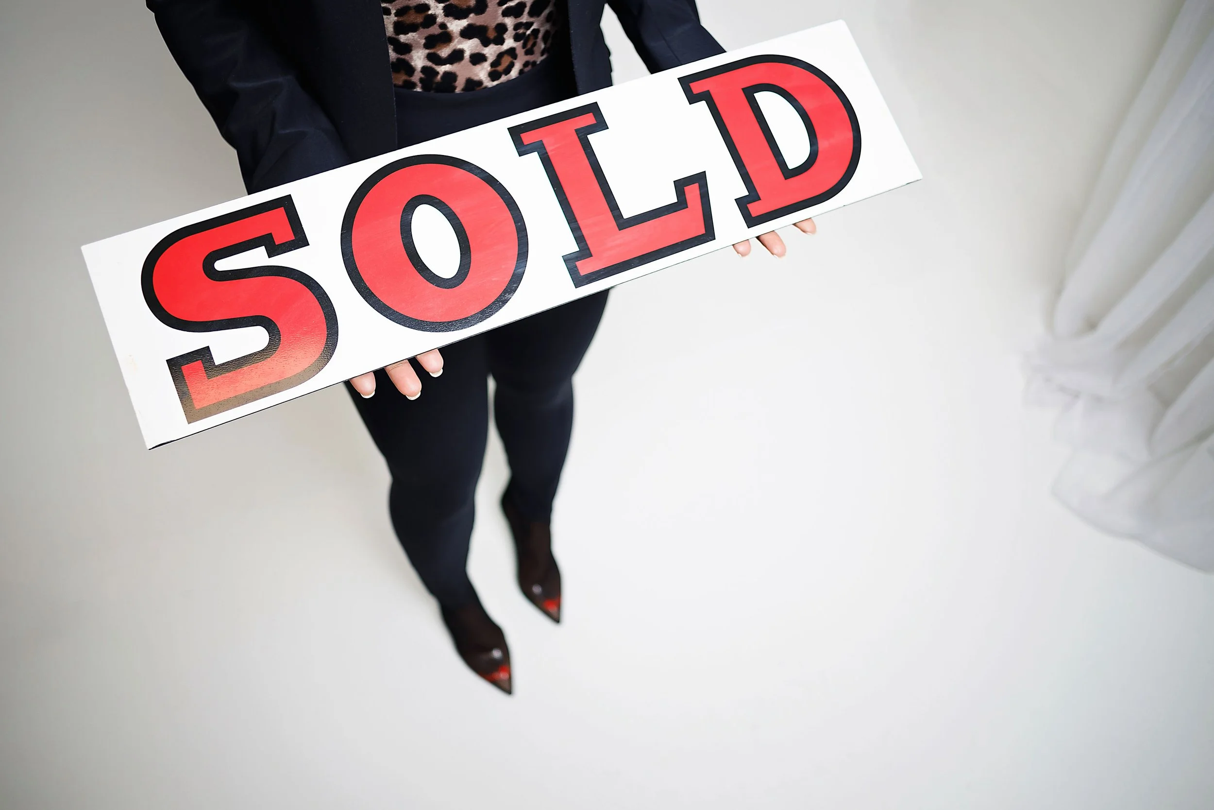 Person holding a white sign with the word "SOLD" in large red and black letters, standing indoors by a window with white curtains.