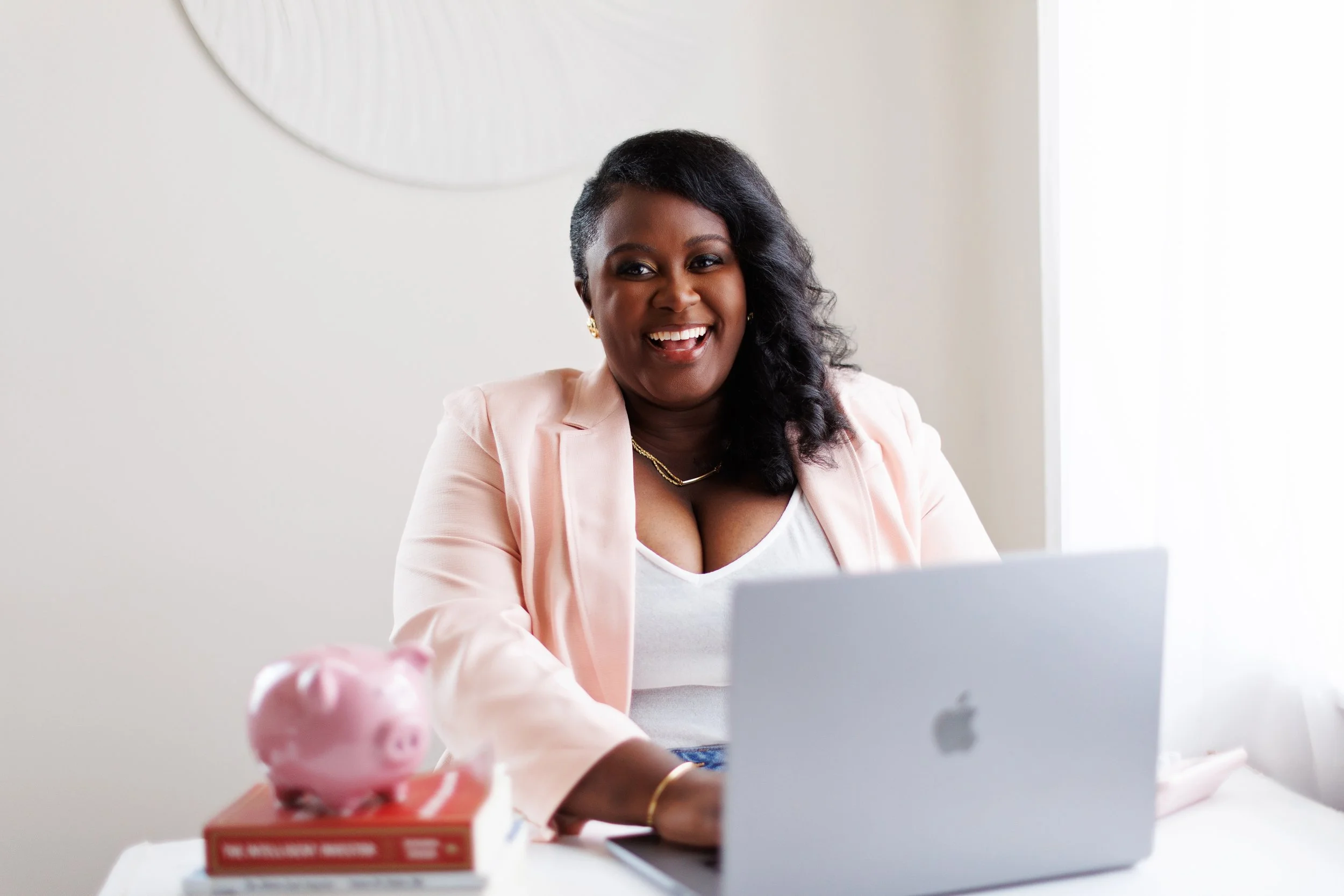 A smiling woman with dark hair in a side-swept style, wearing a light pink blazer and gold jewelry, sitting at a desk with a silver MacBook, pink piggy bank, and a couple of books.
