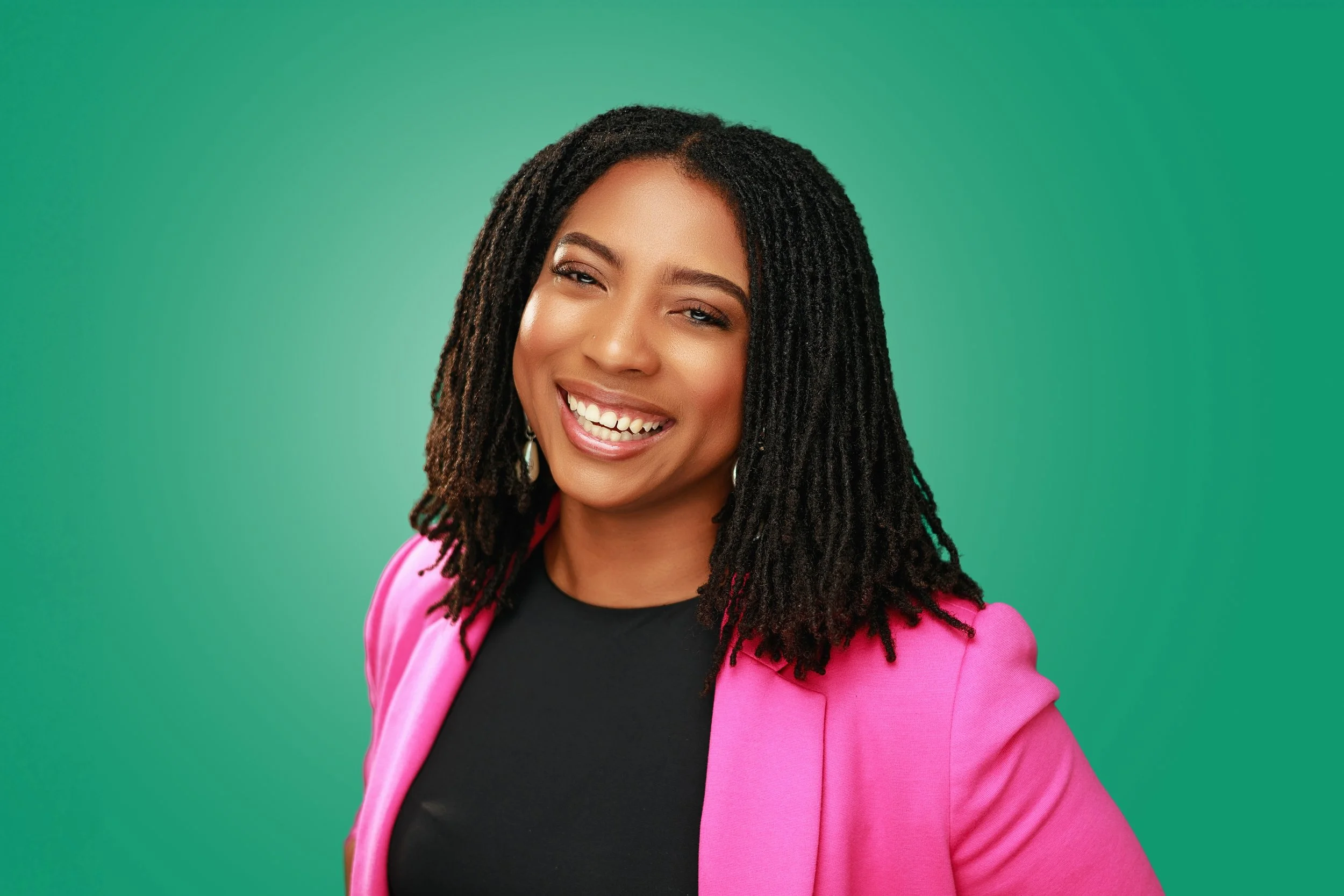 A woman with dark curly hair, wearing a bright pink blazer and black top, smiling against a green background.