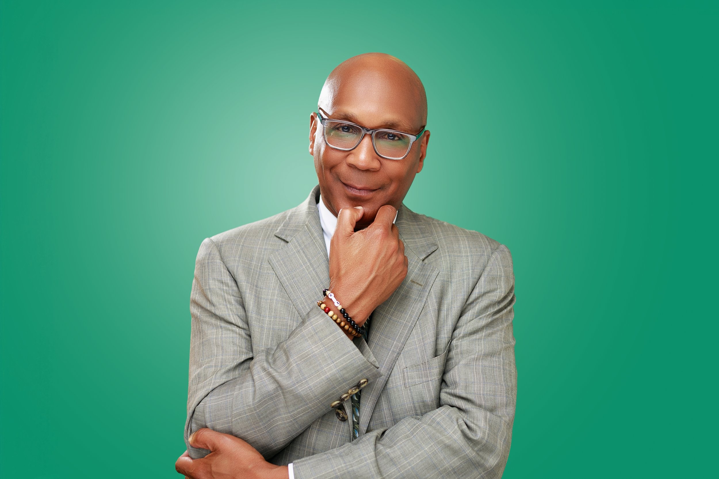 A confident middle-aged man with glasses, wearing a light gray plaid suit and beaded bracelets, standing against a green background.