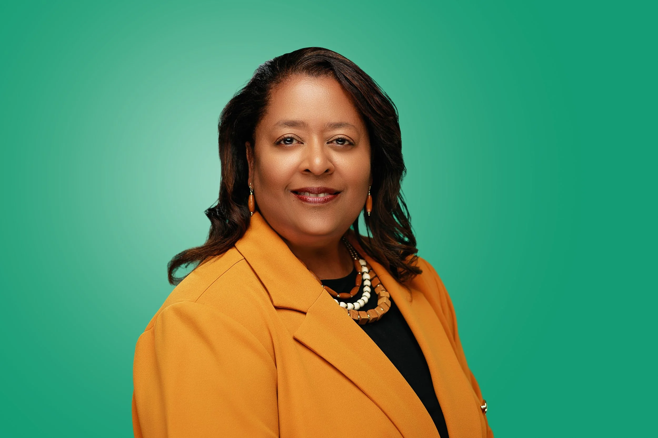 Portrait of an African American woman with shoulder-length wavy hair wearing an orange blazer, black top, and layered necklaces, set against a green background.