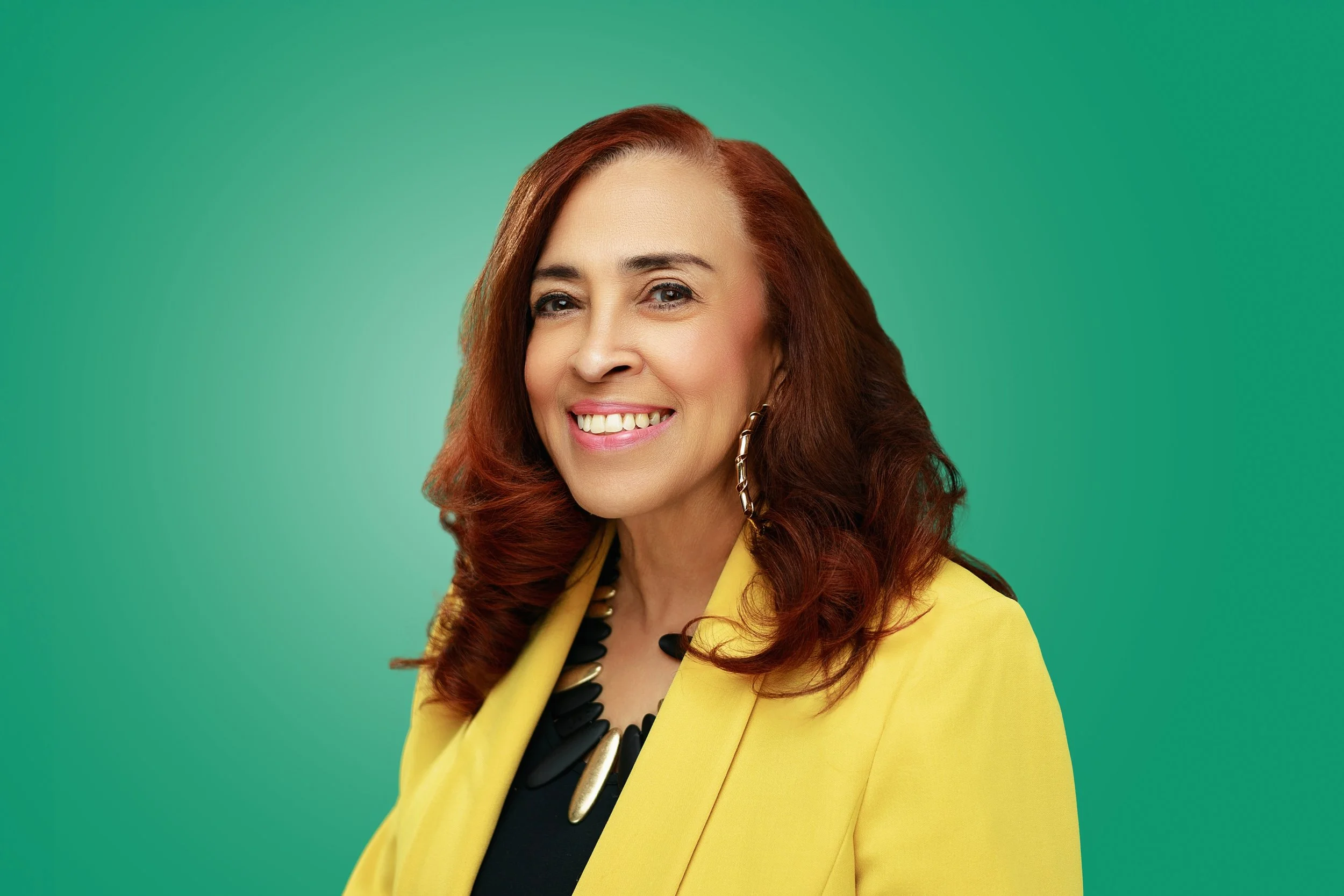 Portrait of a woman with long red hair, smiling, wearing a yellow blazer, black top, and chunky necklace, against a green background.
