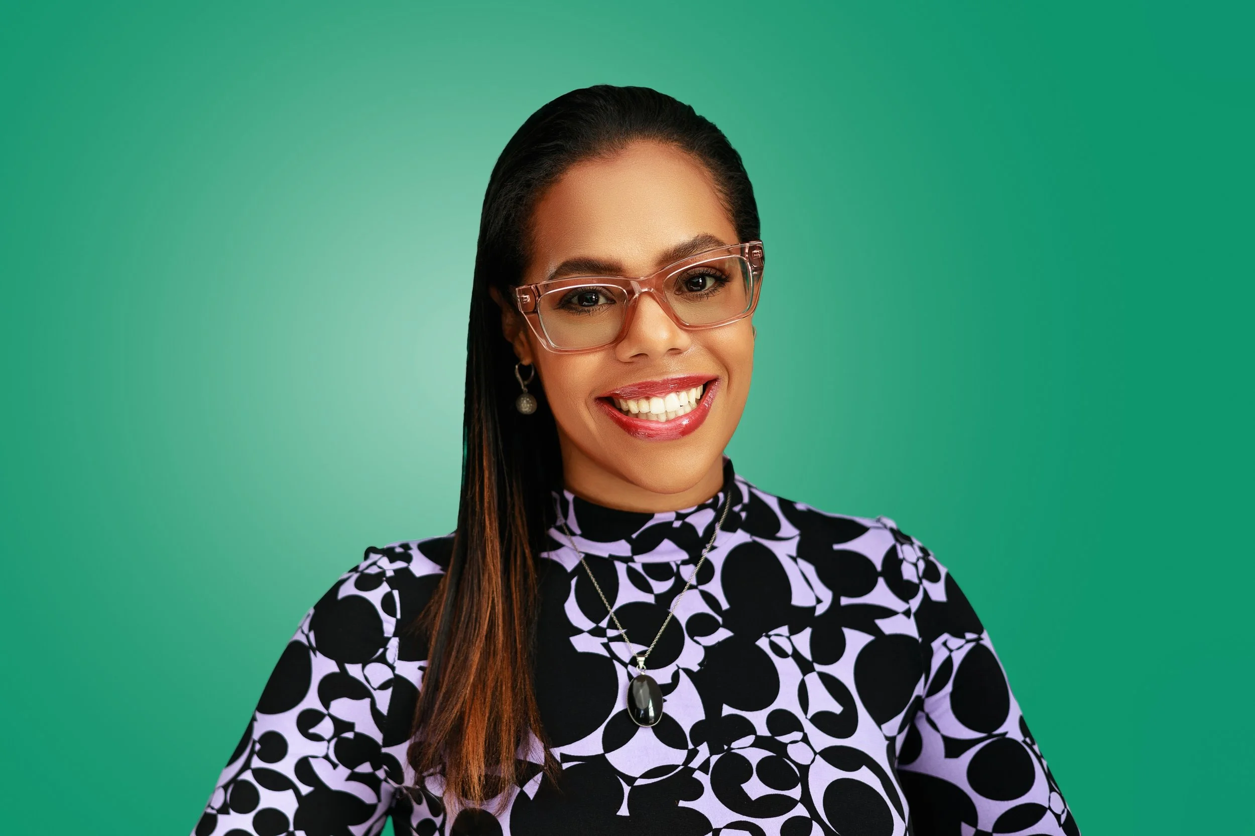 A smiling woman with long dark hair, wearing glasses, a black and white patterned top, and jewelry, against a green background.