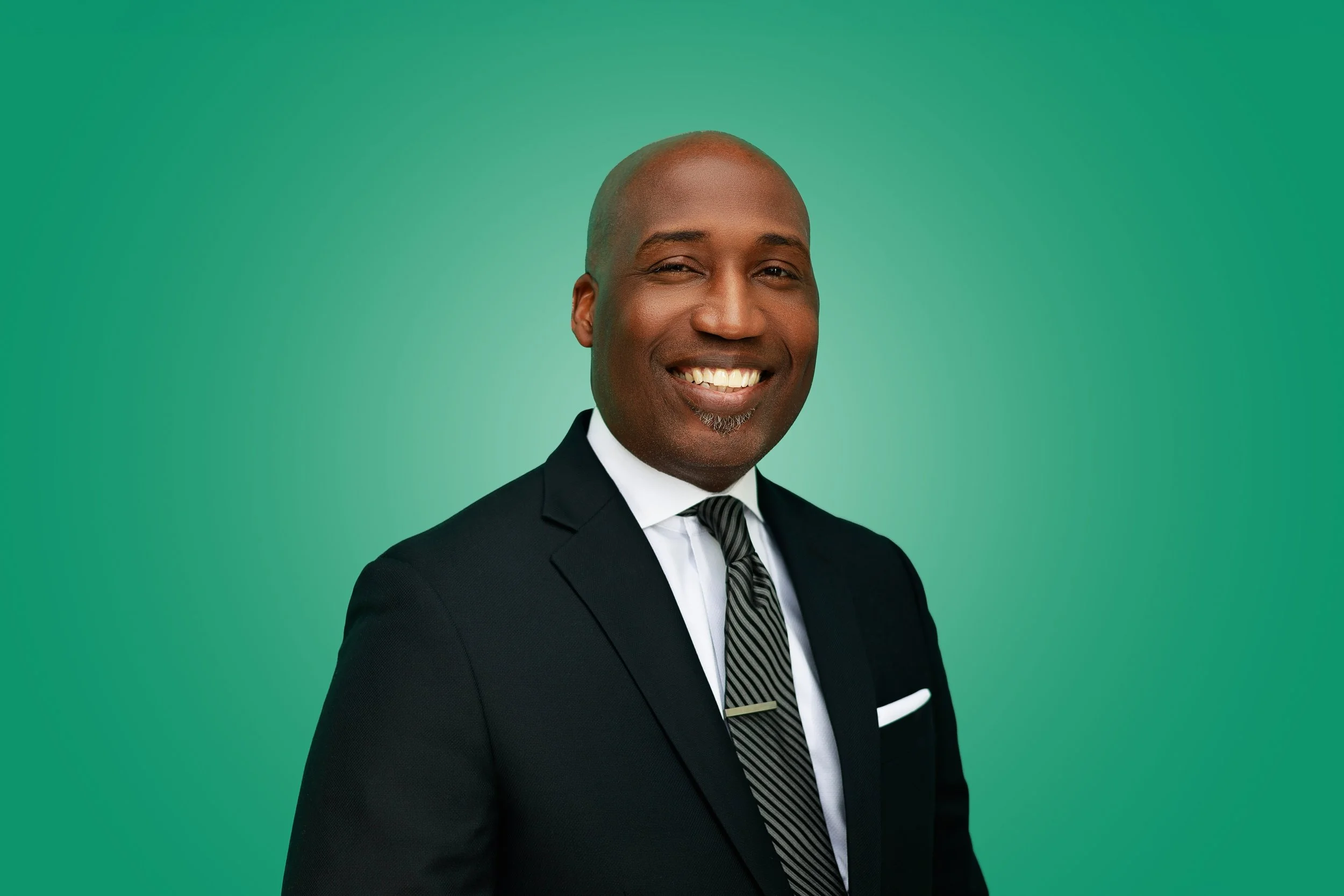 Portrait of a smiling African American man in a formal black suit, white shirt, and striped tie, against a solid green background.