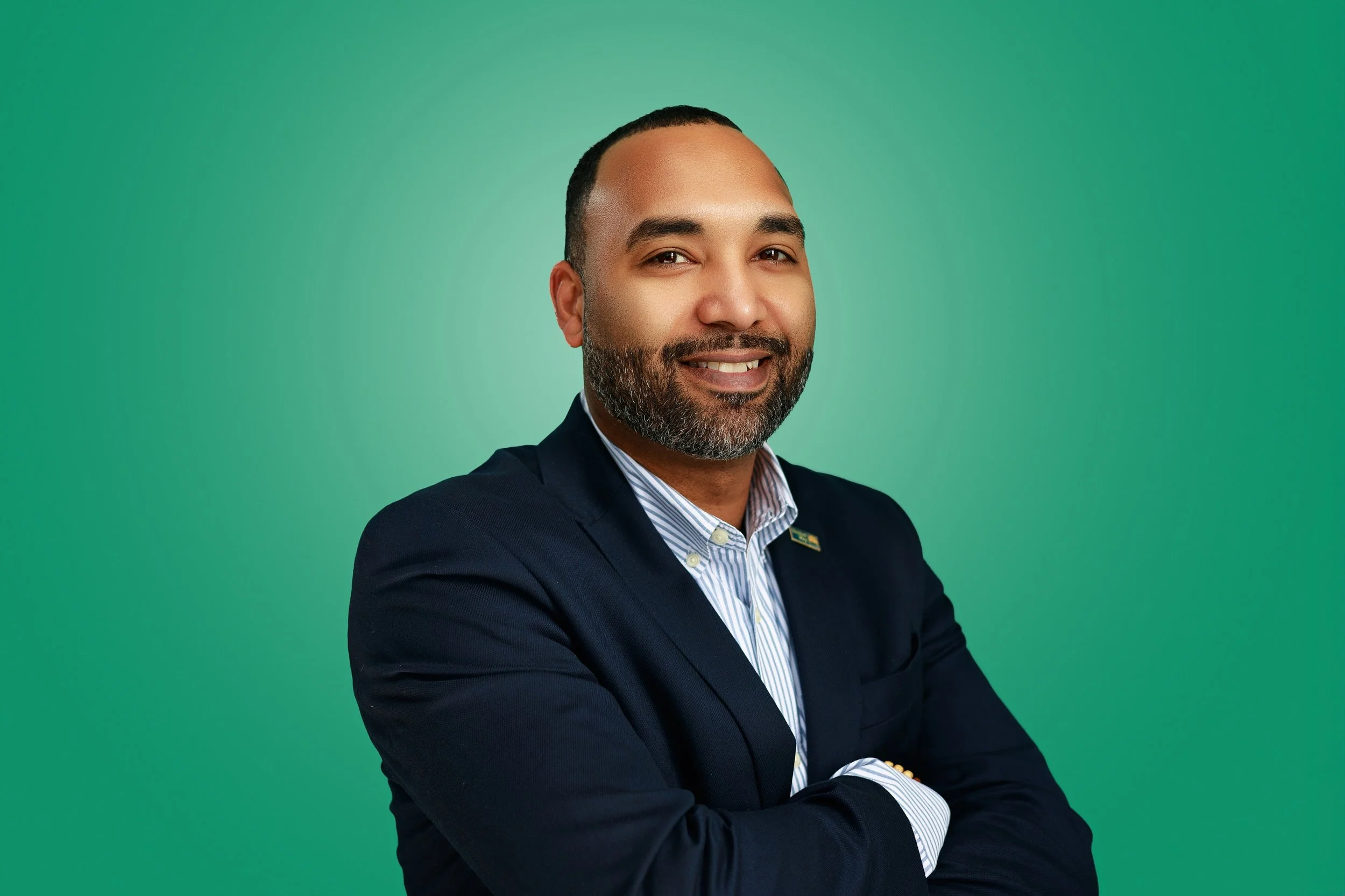 Portrait of a smiling man with a beard in a navy blazer and striped shirt against a green background.