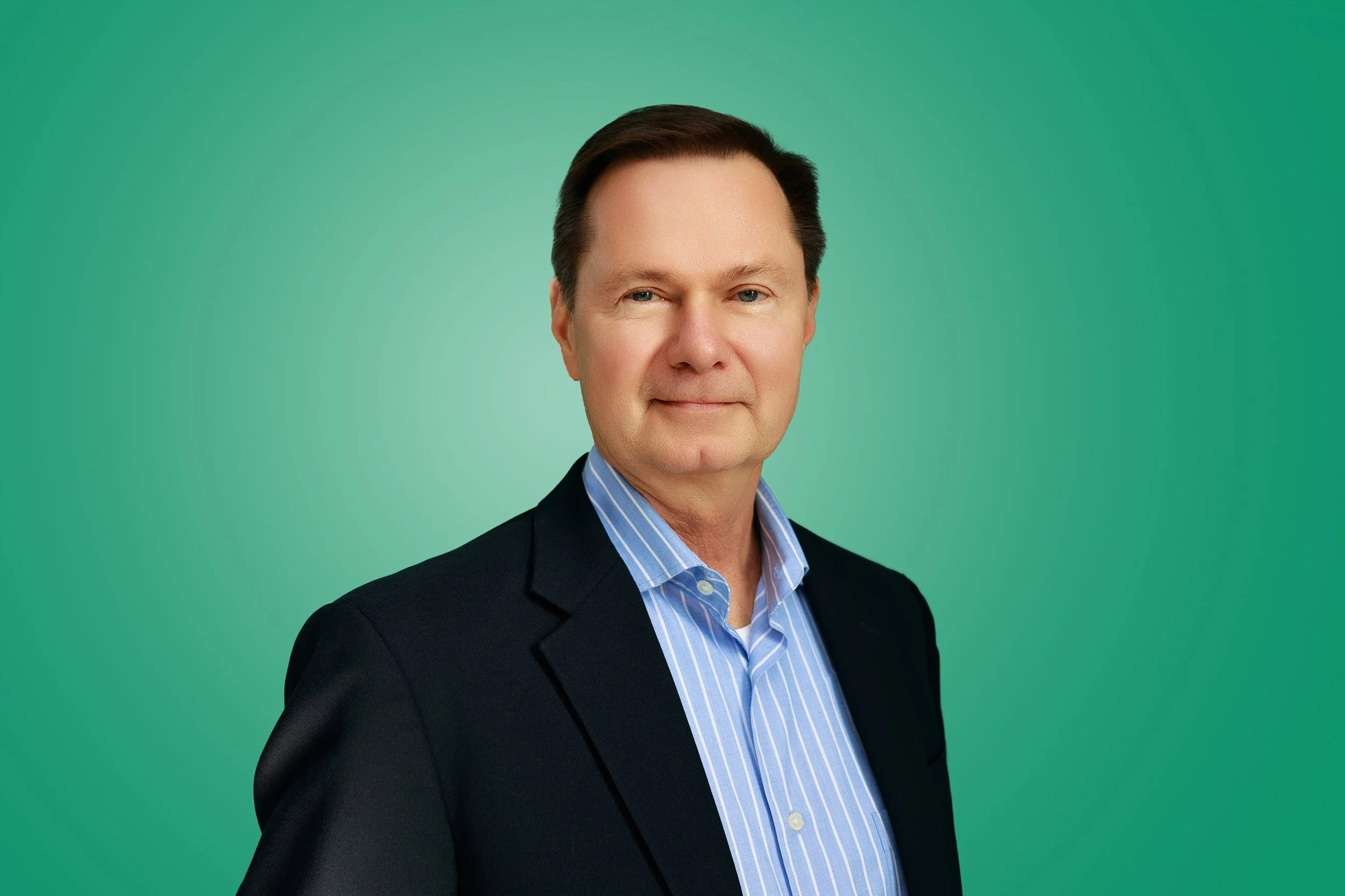 A middle-aged man with dark brown hair, wearing a dark blazer and a blue striped shirt, posing against a green background.