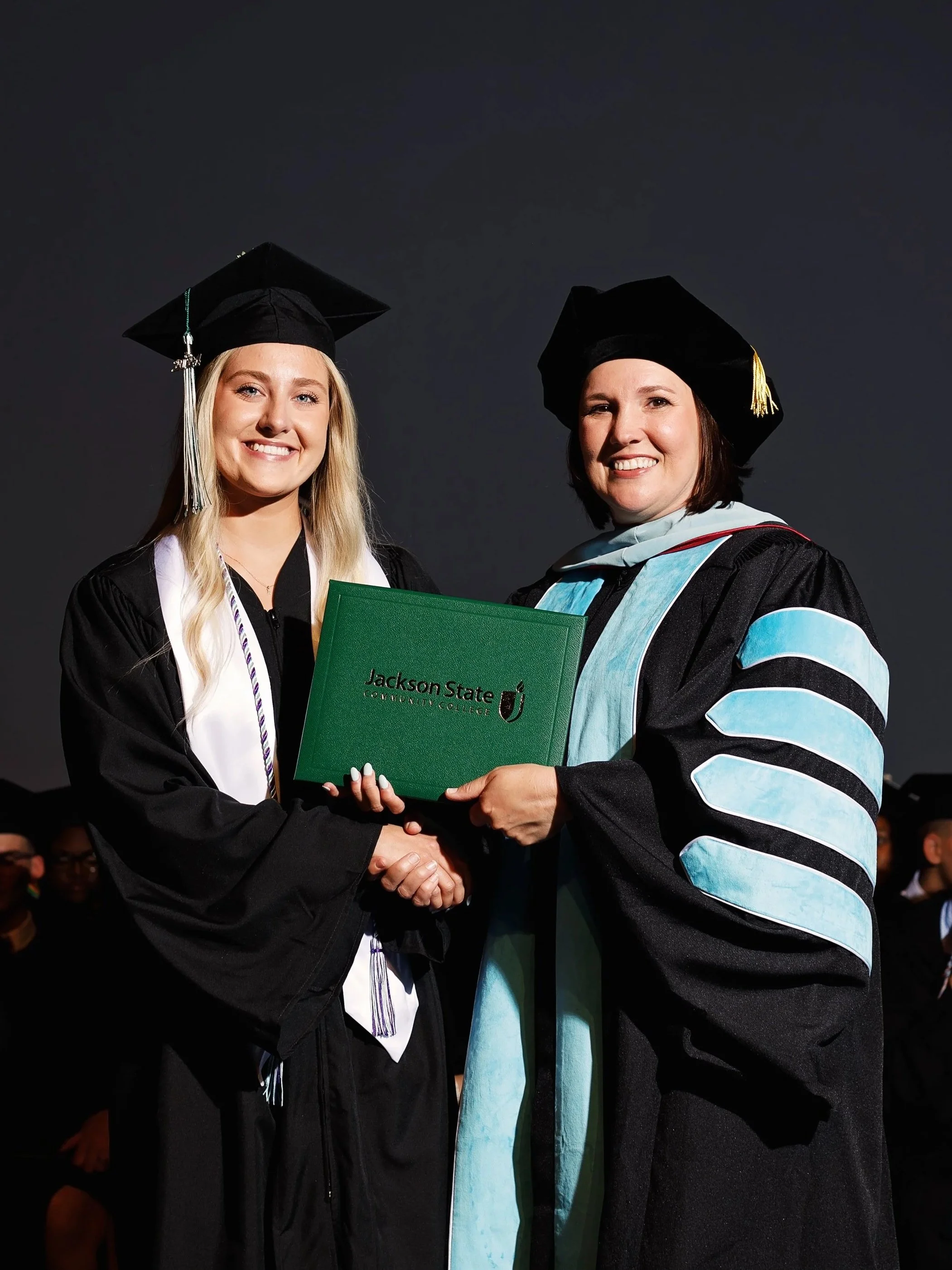 A young woman in a black graduation gown and cap shaking hands and receiving a diploma from a woman in academic regalia at graduation ceremony.