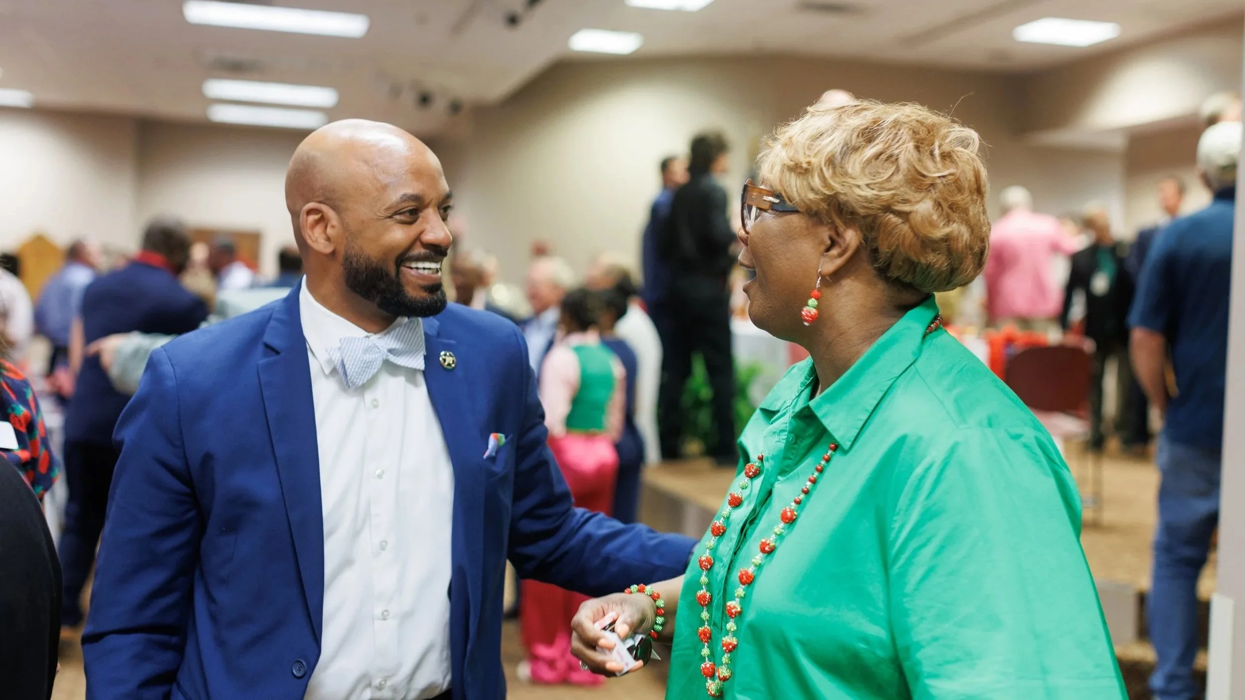 Two people engaged in conversation at a busy indoor event, with one man in a blue suit and bowtie and an older woman in a green shirt, both smiling.