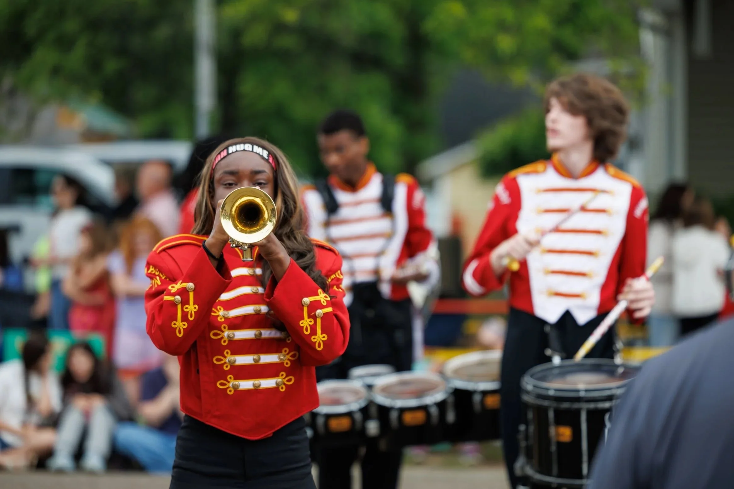 A young female marching band member in a red and gold jacket, playing a trumpet at an outdoor event, with other band members and spectators in the background.