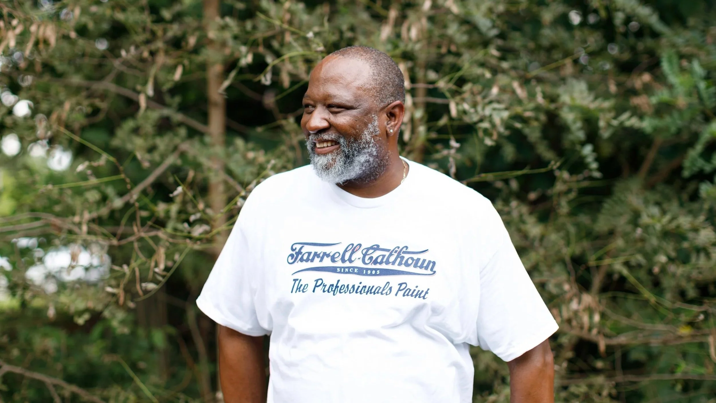 A smiling middle-aged man with a beard and earrings, wearing a white t-shirt with blue text, standing outdoors in front of green foliage.