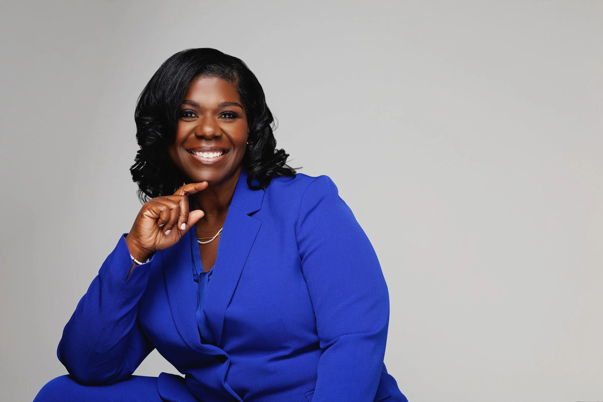 A woman with dark hair smiling, wearing a bright blue blazer, sitting against a plain light gray background.