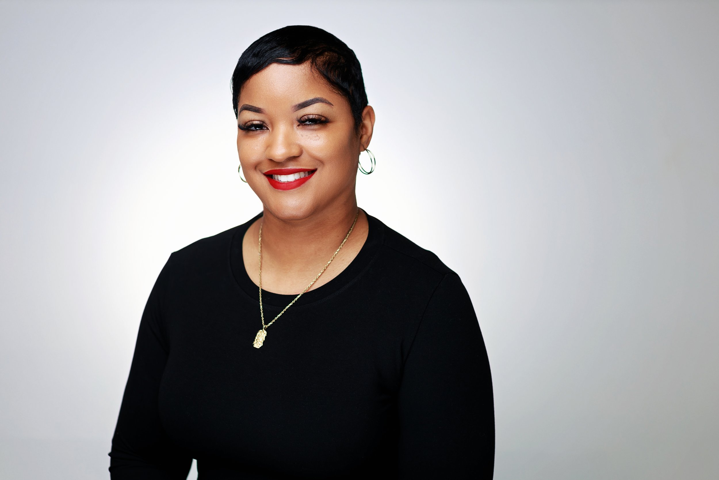 Portrait of a woman with short black hair, red lipstick, wearing a black top, gold necklace, and hoop earrings, smiling against a light gray background.