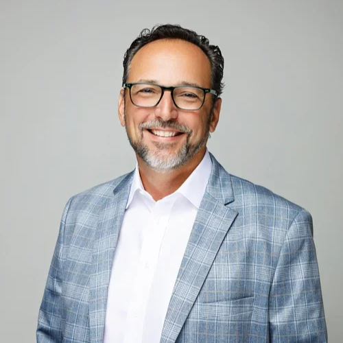 A smiling man with glasses, a gray beard, and dark hair, wearing a light blue checked blazer and a white shirt against a plain gray background.