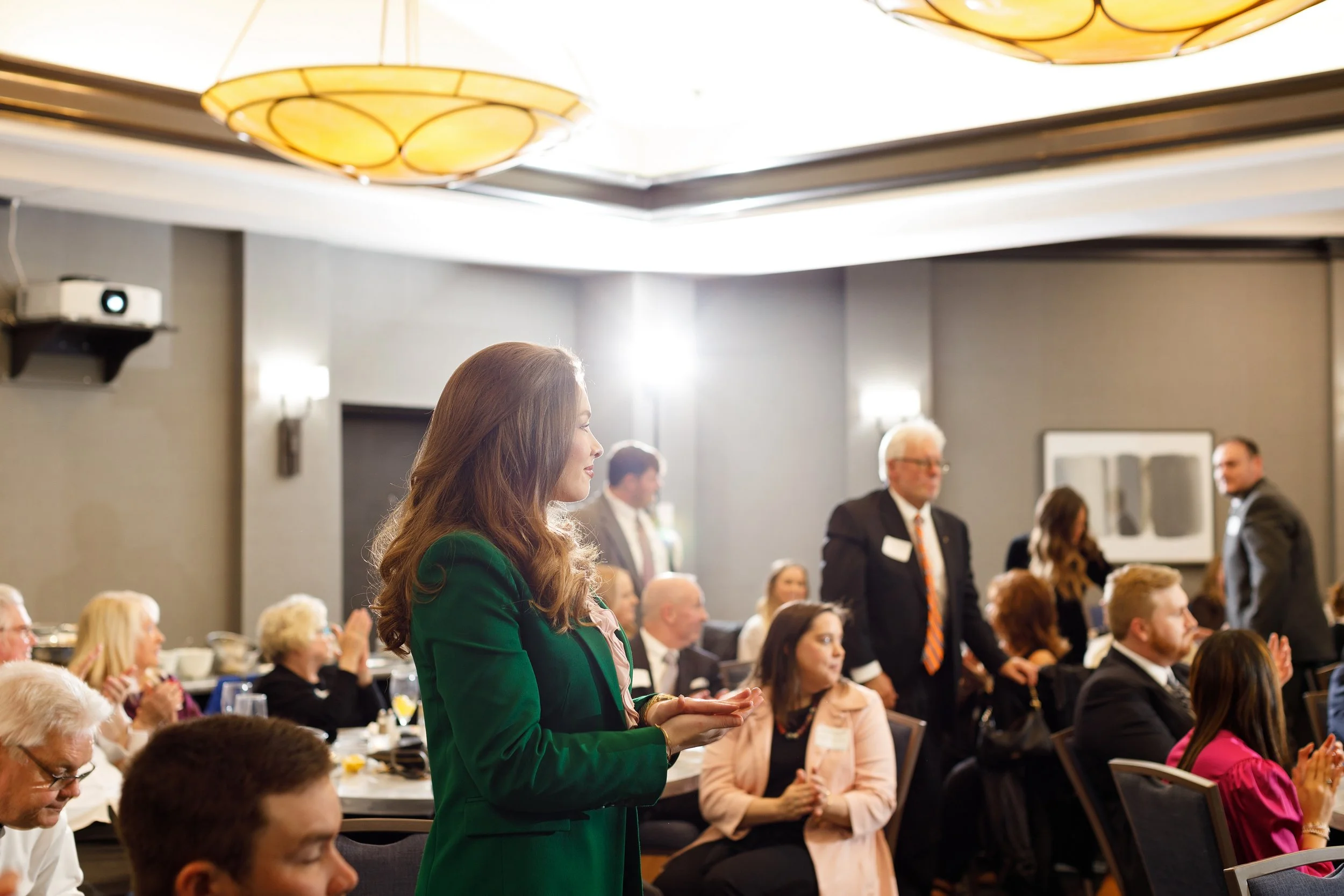 Woman in green blazer standing and clapping at a formal event with seated attendees in a conference room.