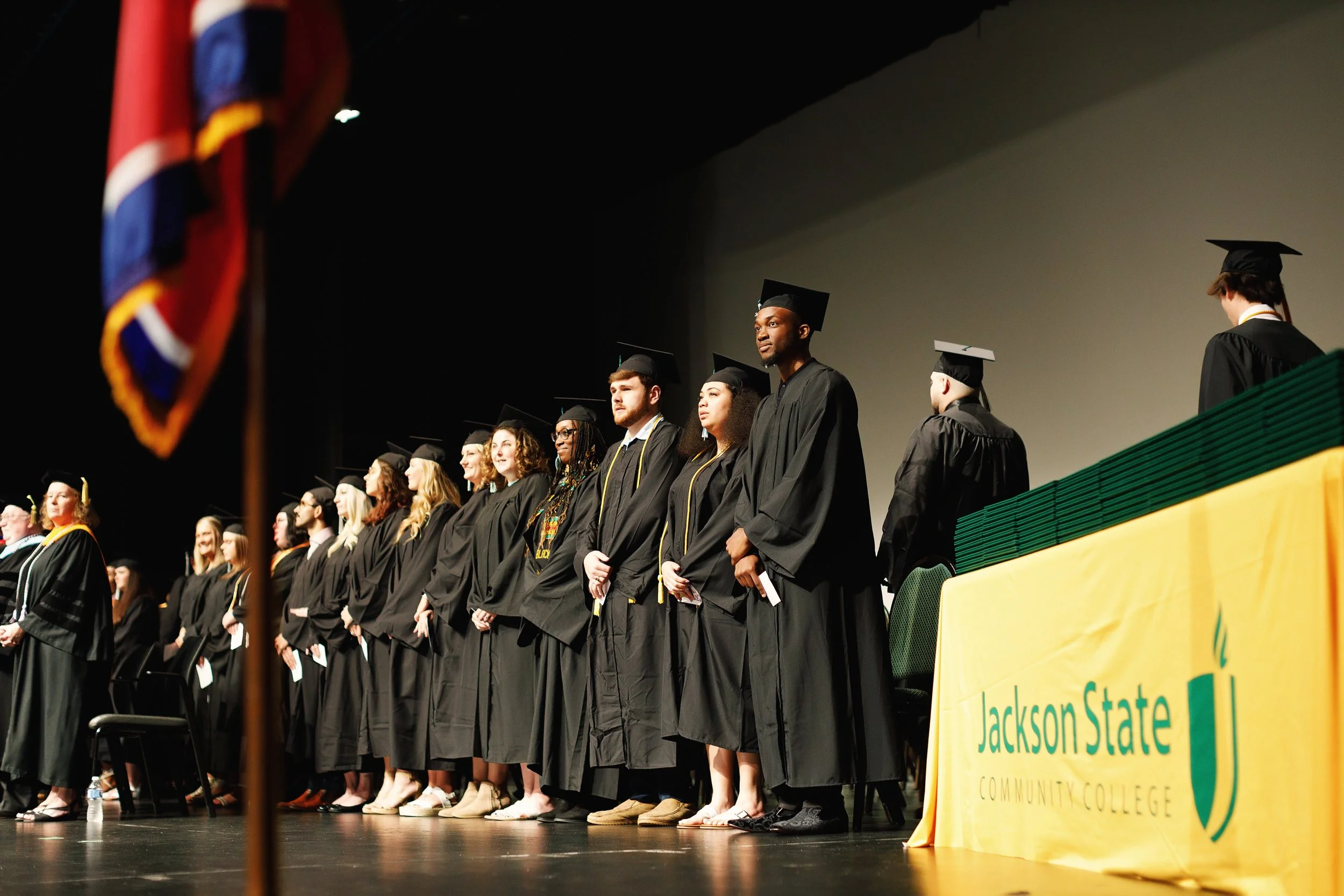 Graduate students in caps and gowns standing on stage at a graduation ceremony, with a banner for Jackson State Community College on the side.
