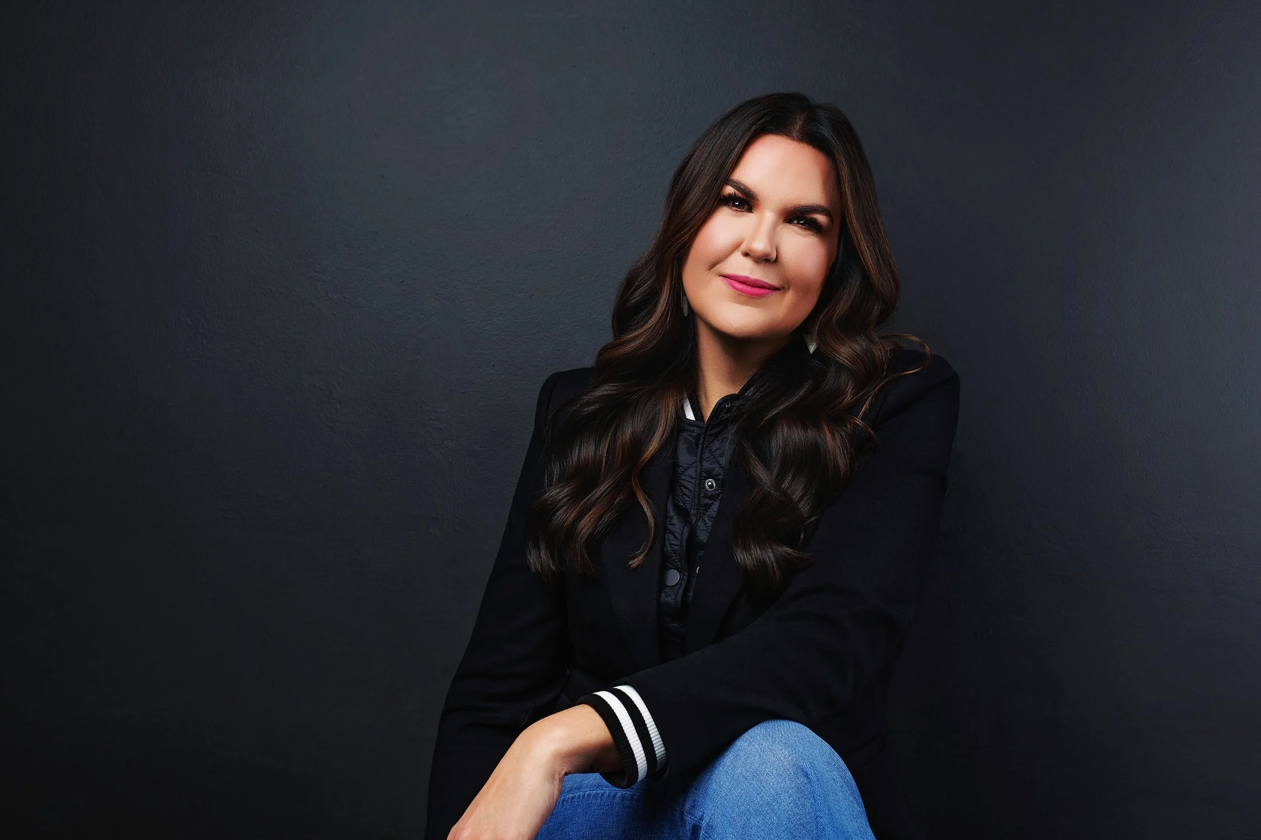 A woman with long brown wavy hair, wearing a black jacket with white trim, sits against a dark grey textured wall. She has pink lipstick and a subtle smile, with one arm resting on her knee.