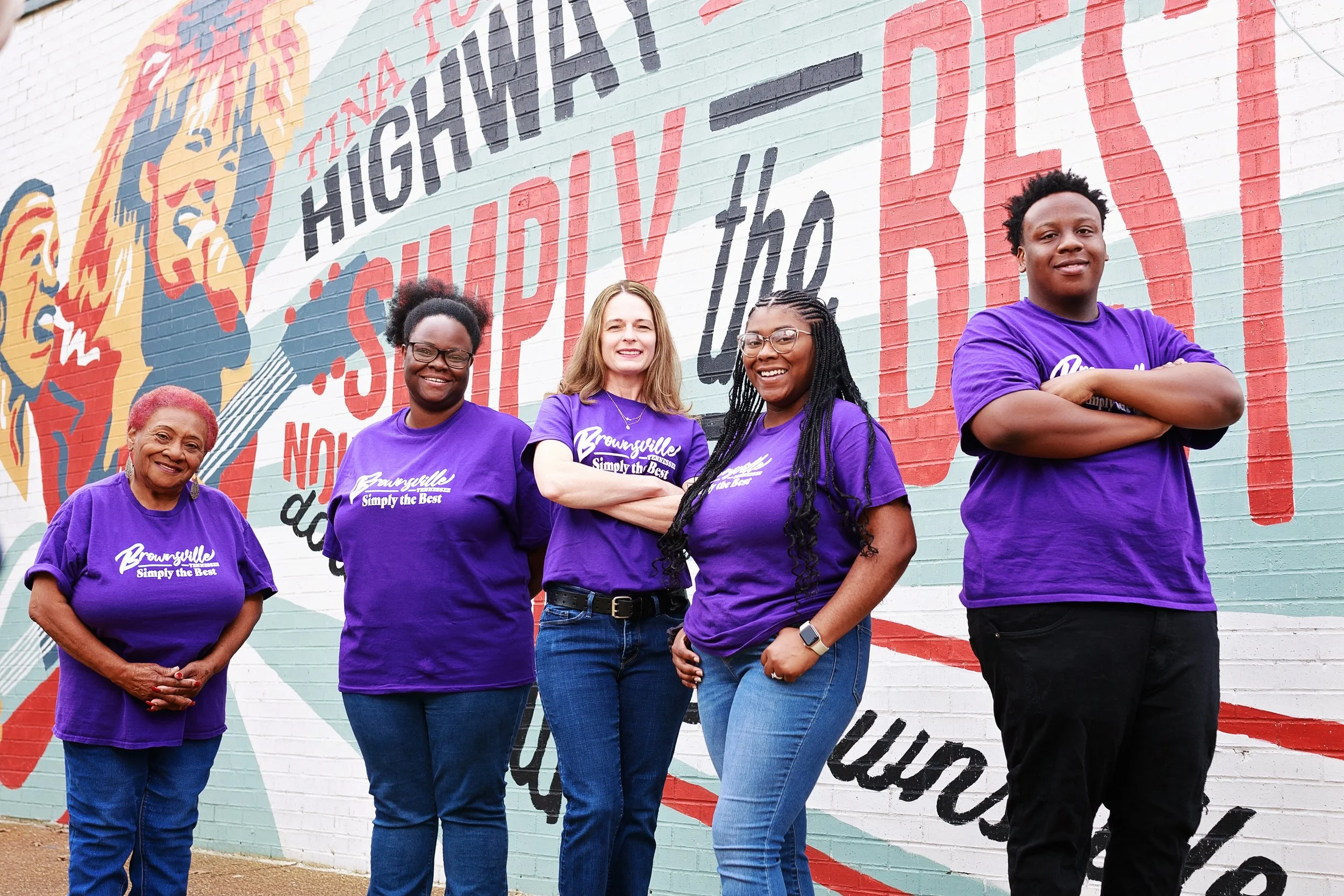 Five women standing side by side in front of a colorful mural wall, all wearing purple T-shirts that say 'Brownsville Simply the Best,' smiling at the camera.
