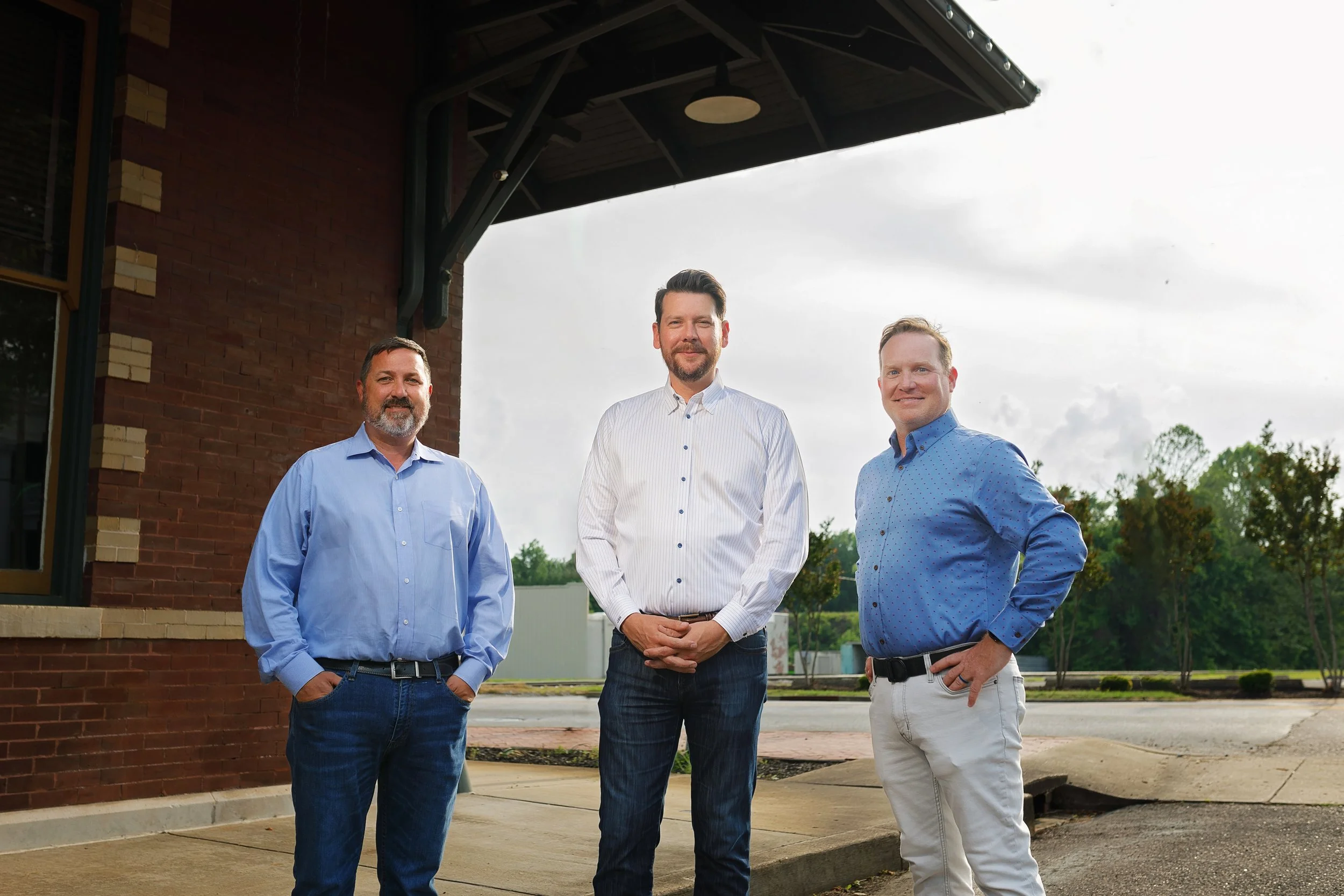 Three men standing outdoors in front of a brick building, dressed in casual business attire, with trees and cloudy sky in the background.