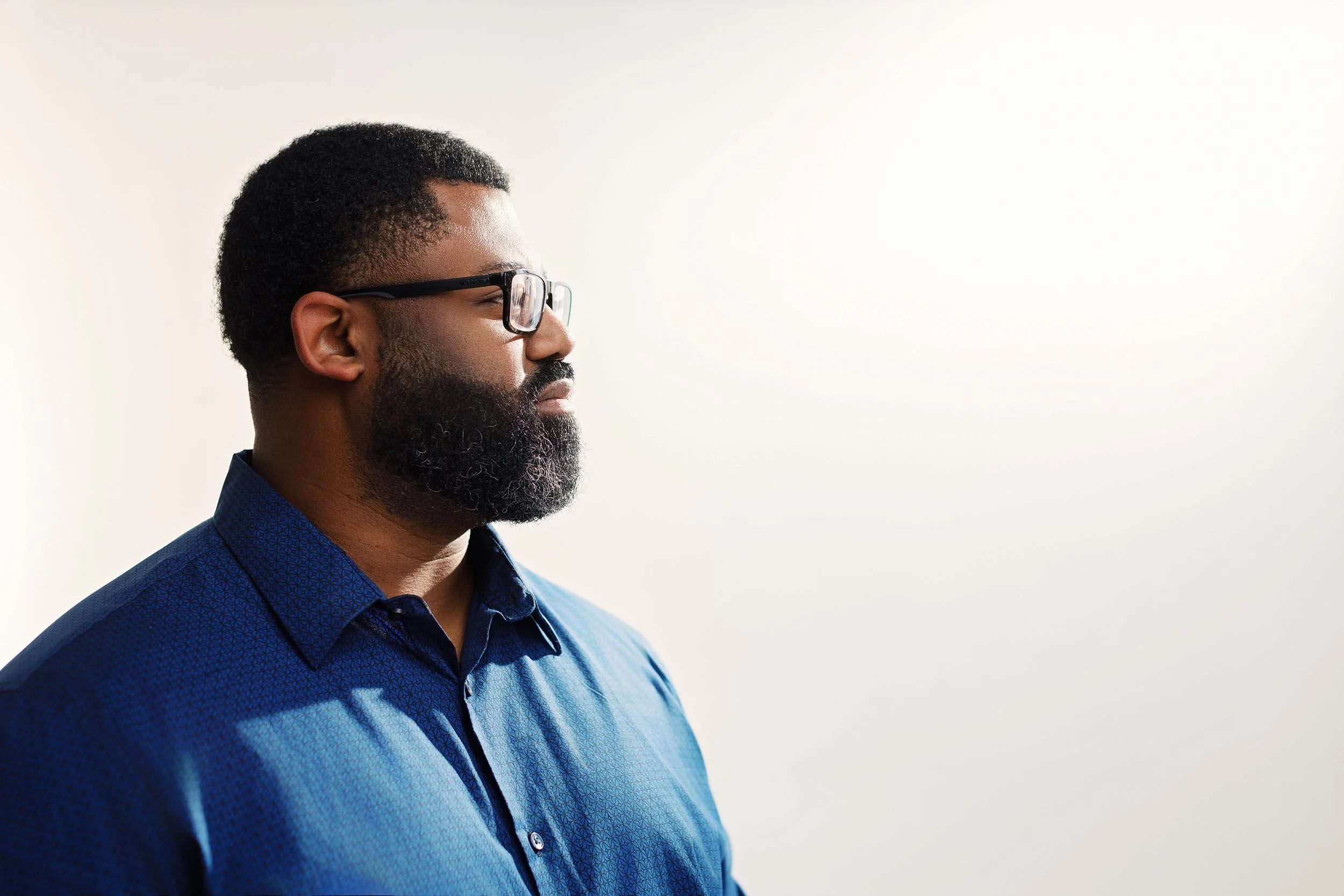 Side view of a man with glasses and a beard wearing a blue shirt against a white background.