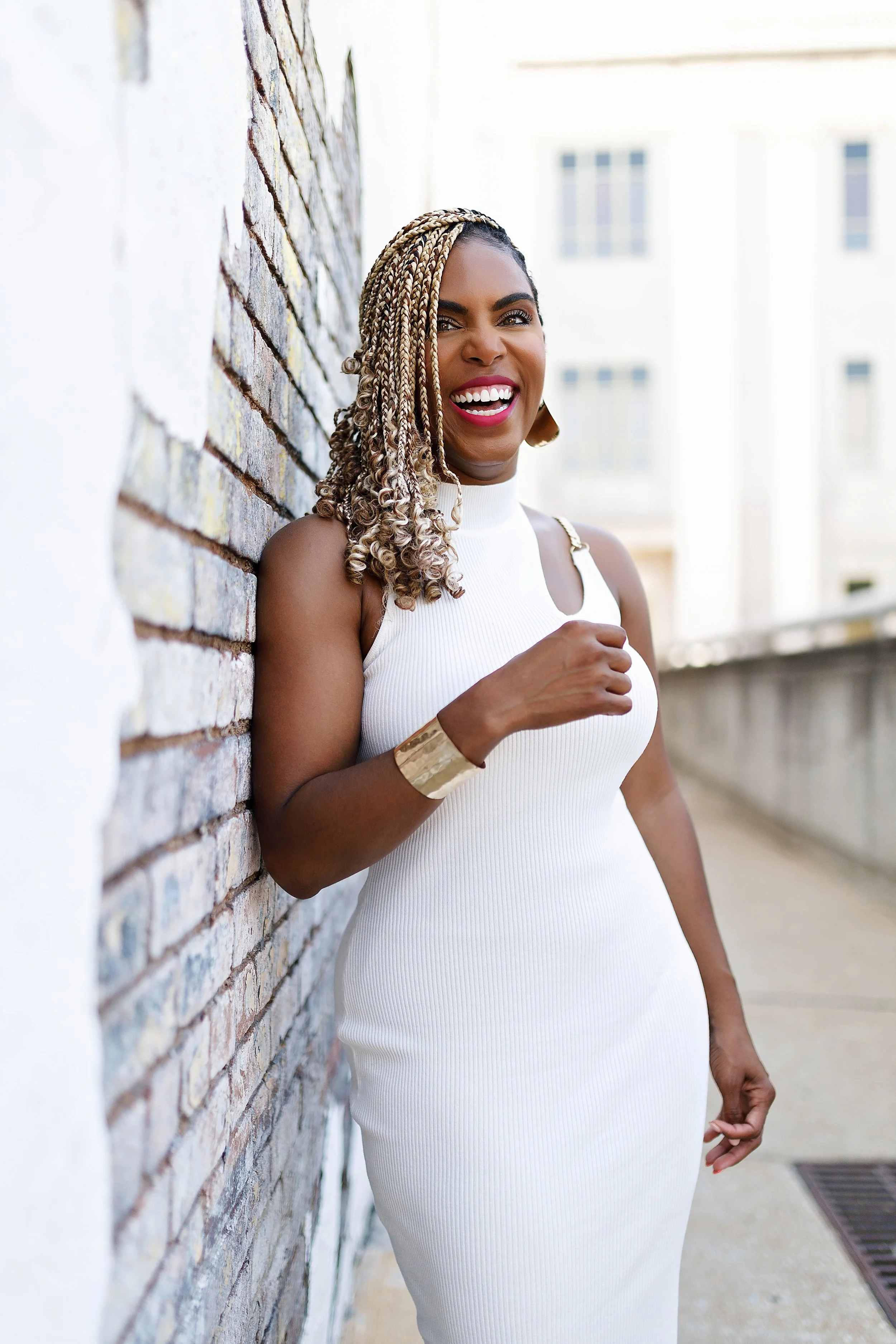 A woman with braided hair in blonde and dark tones, wearing a white sleeveless dress and gold accessories, leaning against a brick wall outside and smiling.