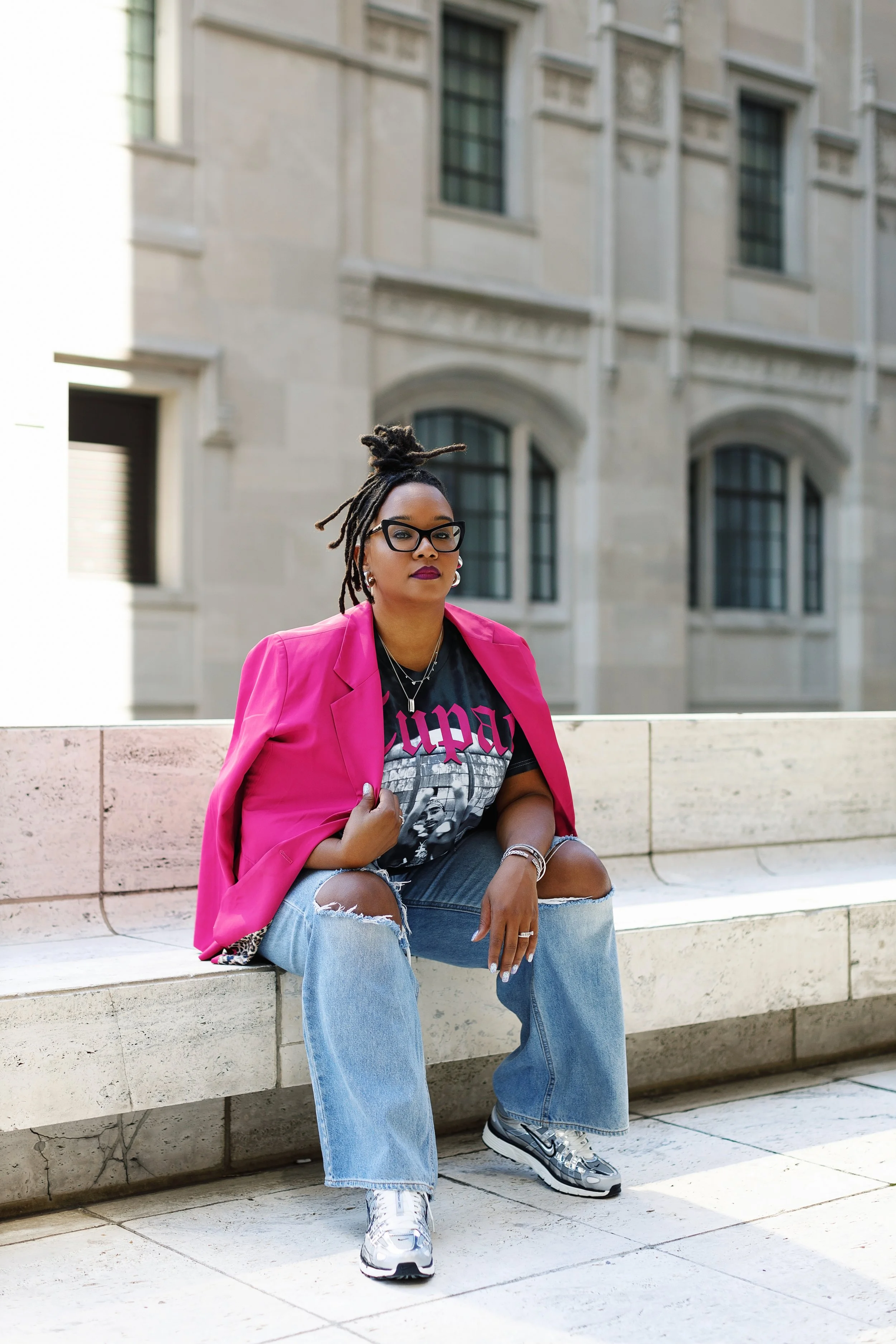 A woman with dreadlocks, glasses, dark lipstick, wearing a pink blazer draped over her shoulders, a black graphic T-shirt, ripped jeans, and sneakers, sitting on a stone bench outside an urban stone building.