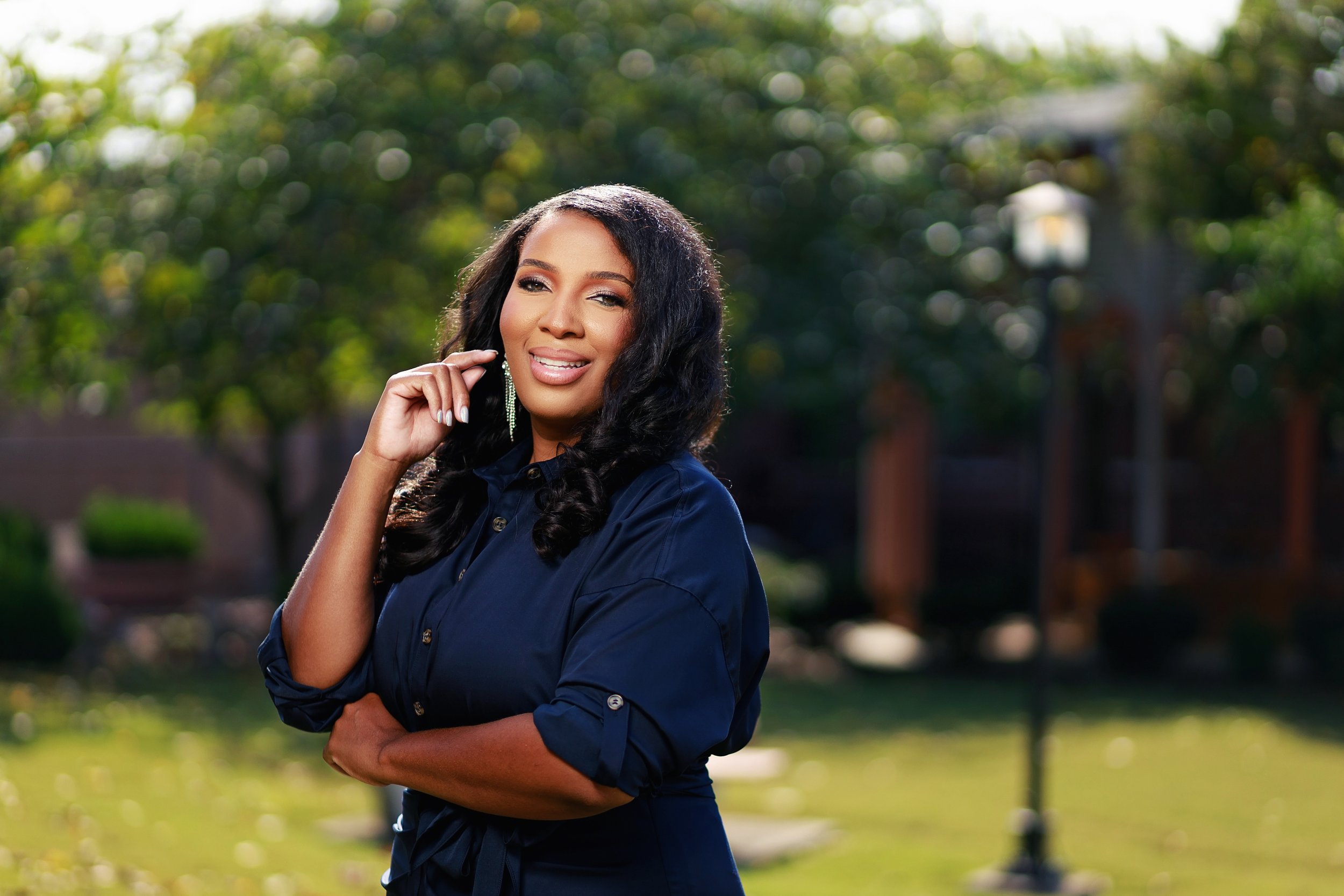 A woman with dark, curly hair wearing a navy blue shirt posing outdoors in a park, smiling with one hand touching her face, blurred trees and a lamp post in the background.