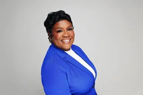 A woman with short curly black hair, wearing a blue blazer and a white shirt, smiling in front of a plain gray background.