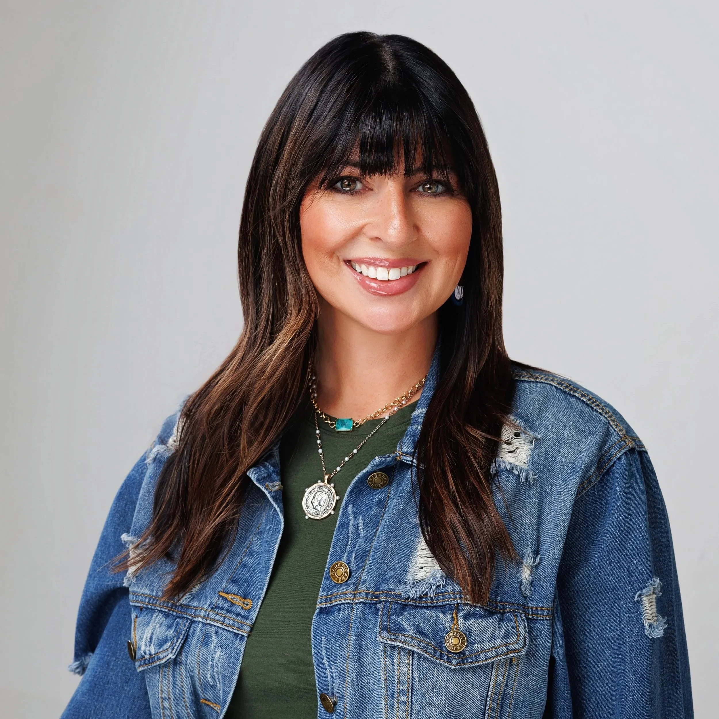 A woman with medium-length brown hair and bangs, smiling, wearing a denim jacket over a green top, and layered necklaces.