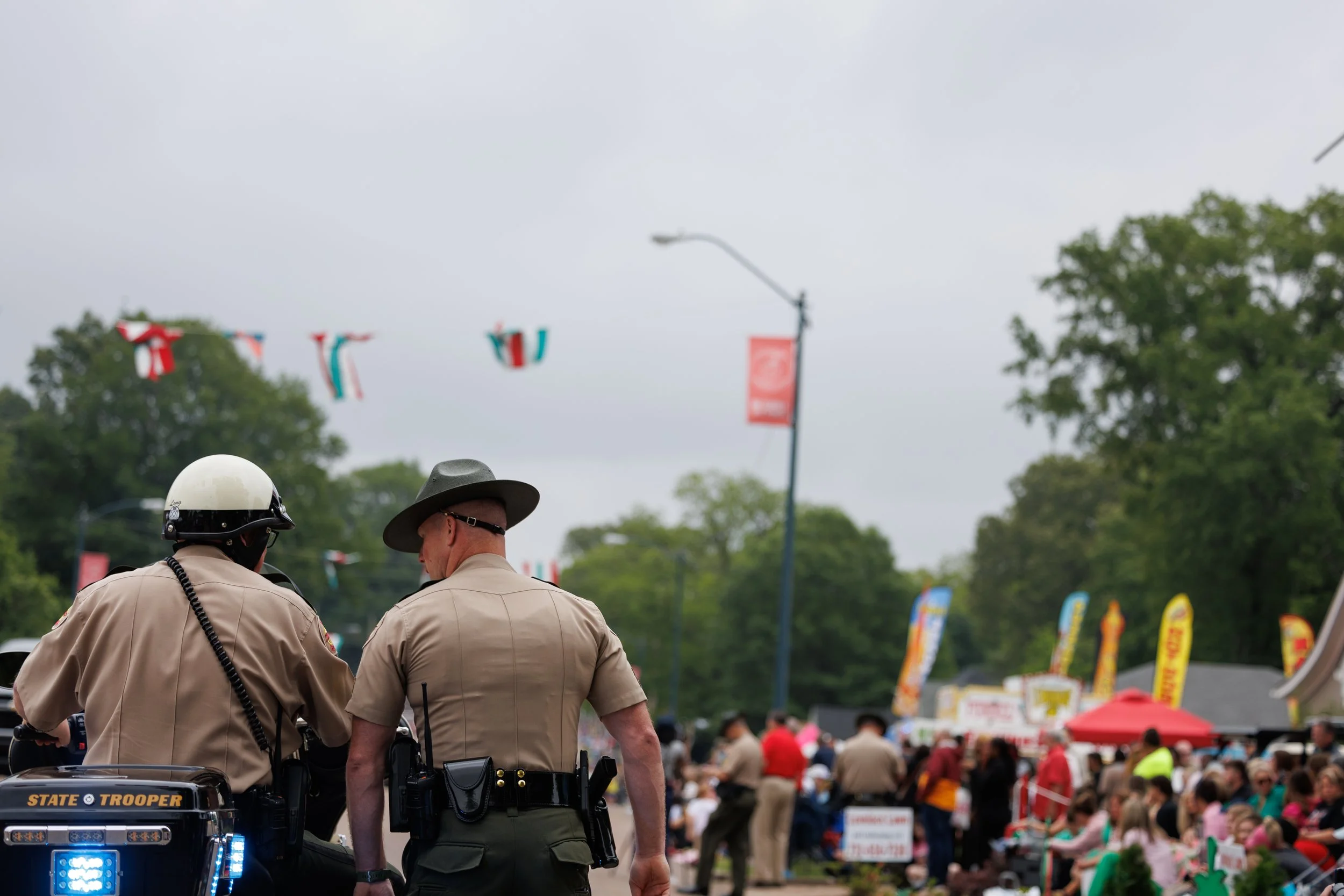 Two uniformed police officers, one wearing a helmet, talking at a crowded outdoor event with tents, flags, and people.