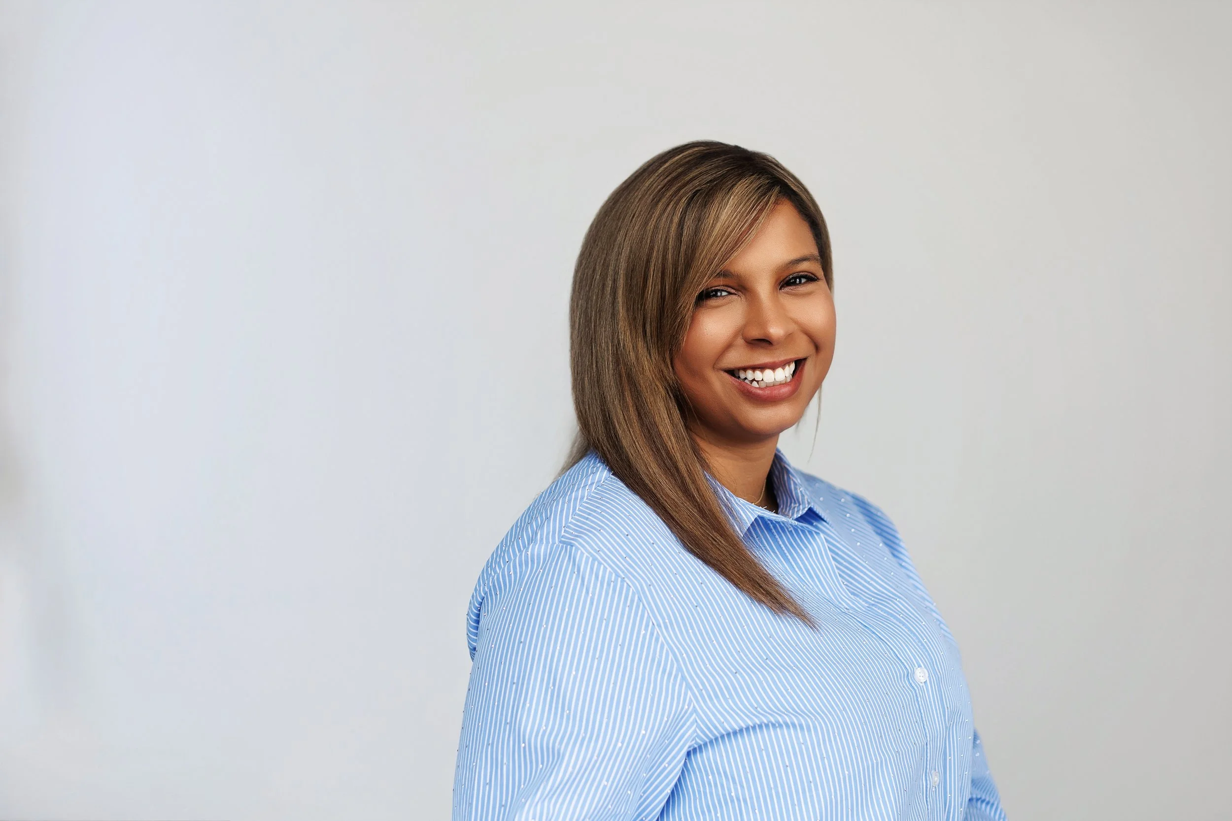 A smiling woman with medium-length brown hair wearing a blue and white striped shirt, standing against a plain white wall.