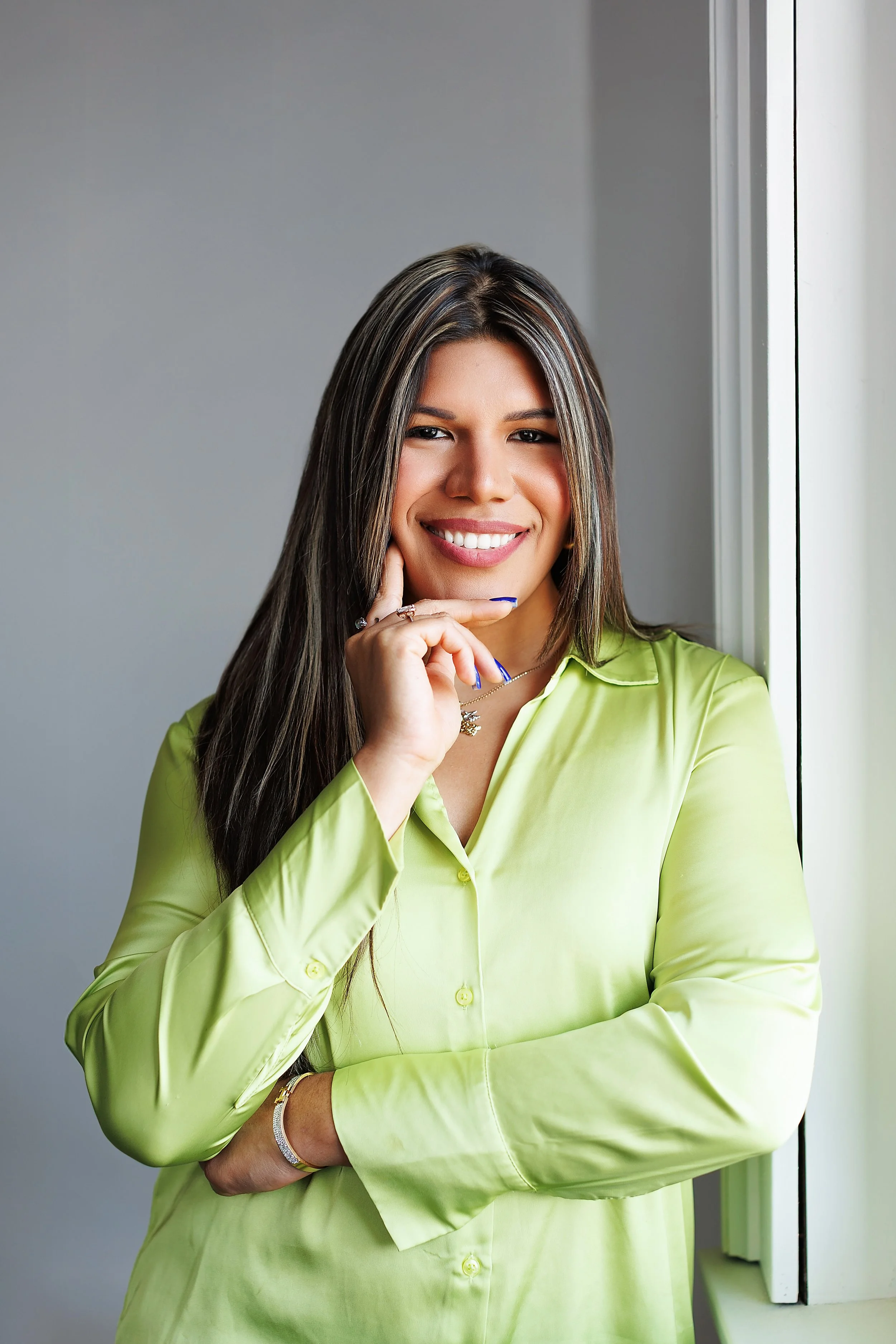 A woman with long dark hair smiling, wearing a light green shirt, standing near a window.