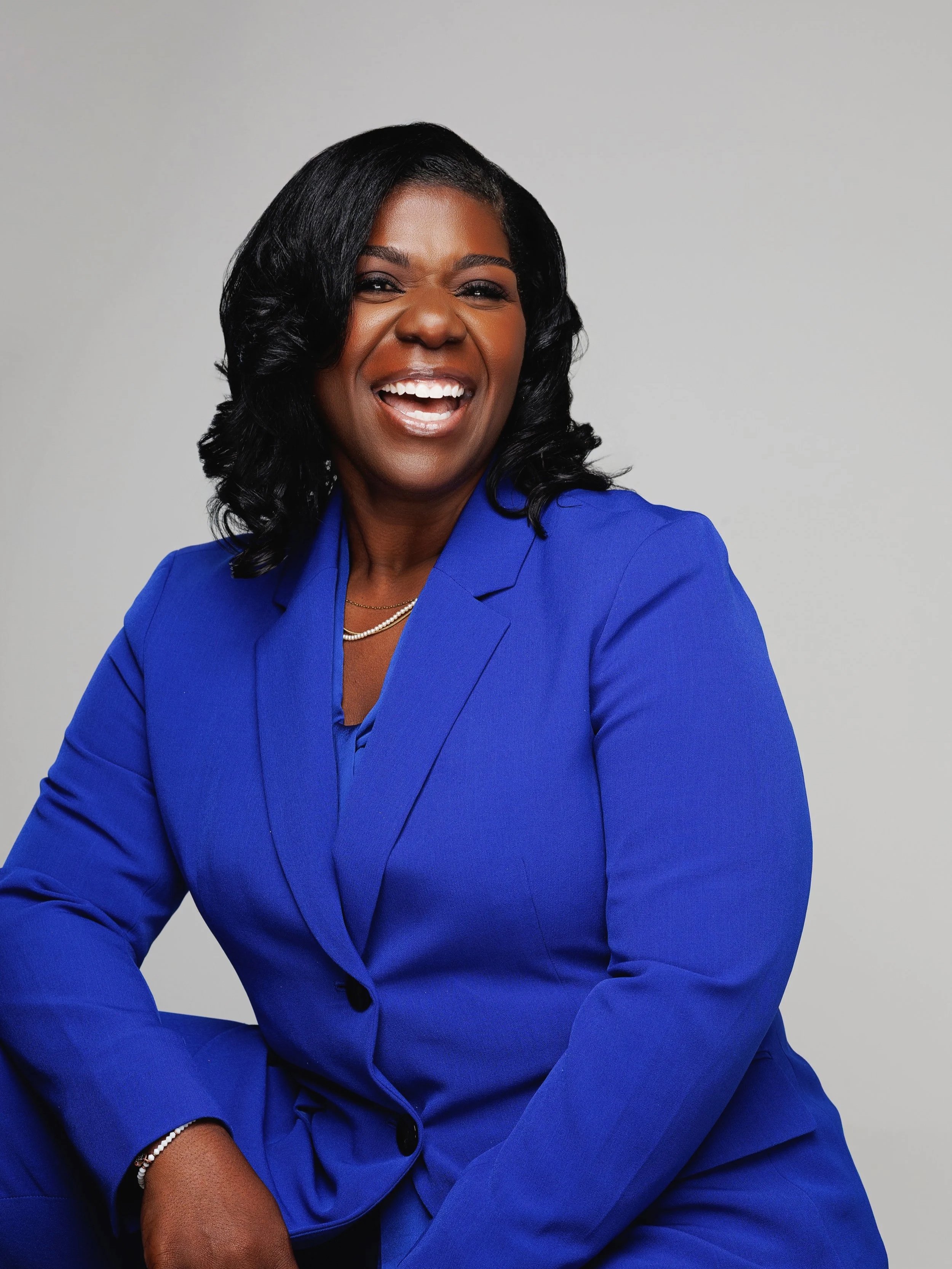 A woman with dark hair wearing a royal blue suit, smiling confidently, against a plain gray background.
