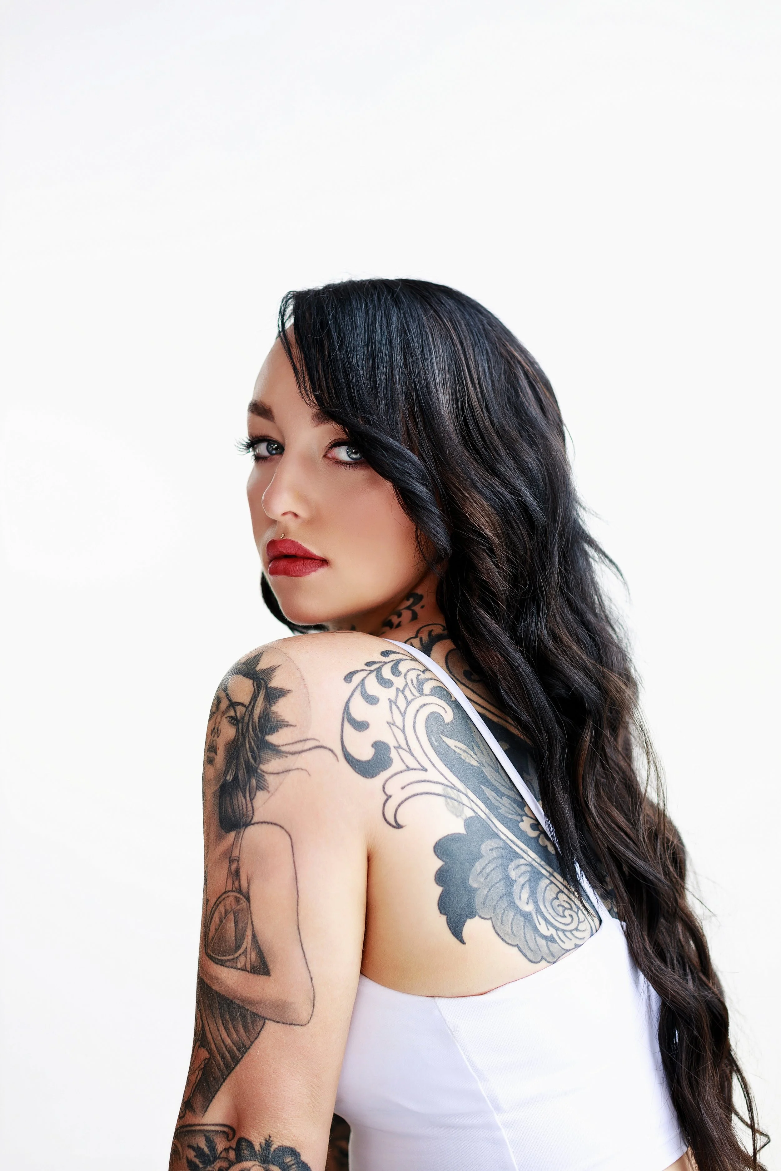 Portrait of a woman with dark wavy hair and tattoos, wearing a white top, looking over her shoulder at the camera.