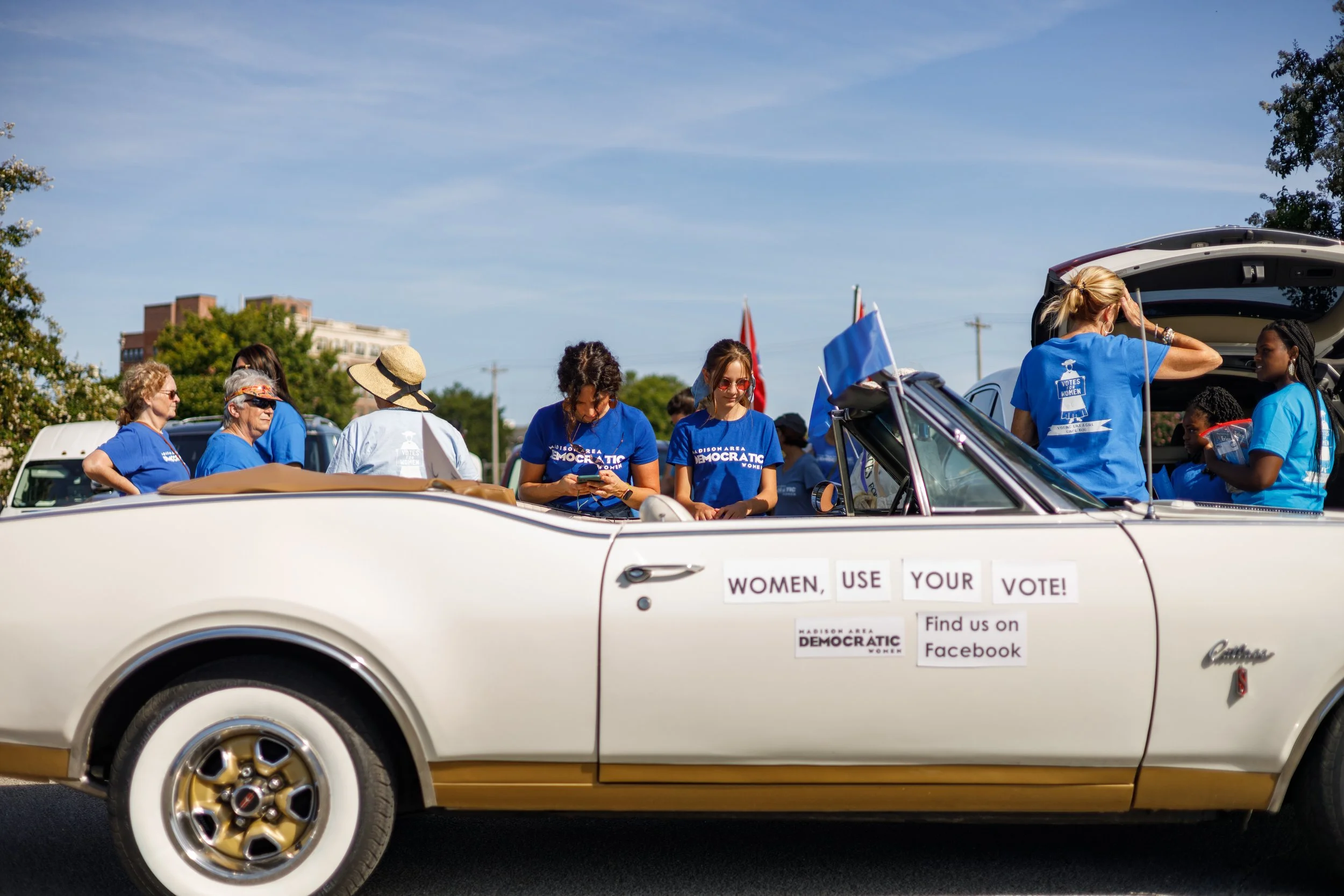 A group of women wearing blue Democratic shirts sitting in and around a vintage white convertible car with signs that say 'Women, Use Your Vote!' and 'Find us on Facebook' during a parade or rally on a sunny day, with trees and buildings in the backg