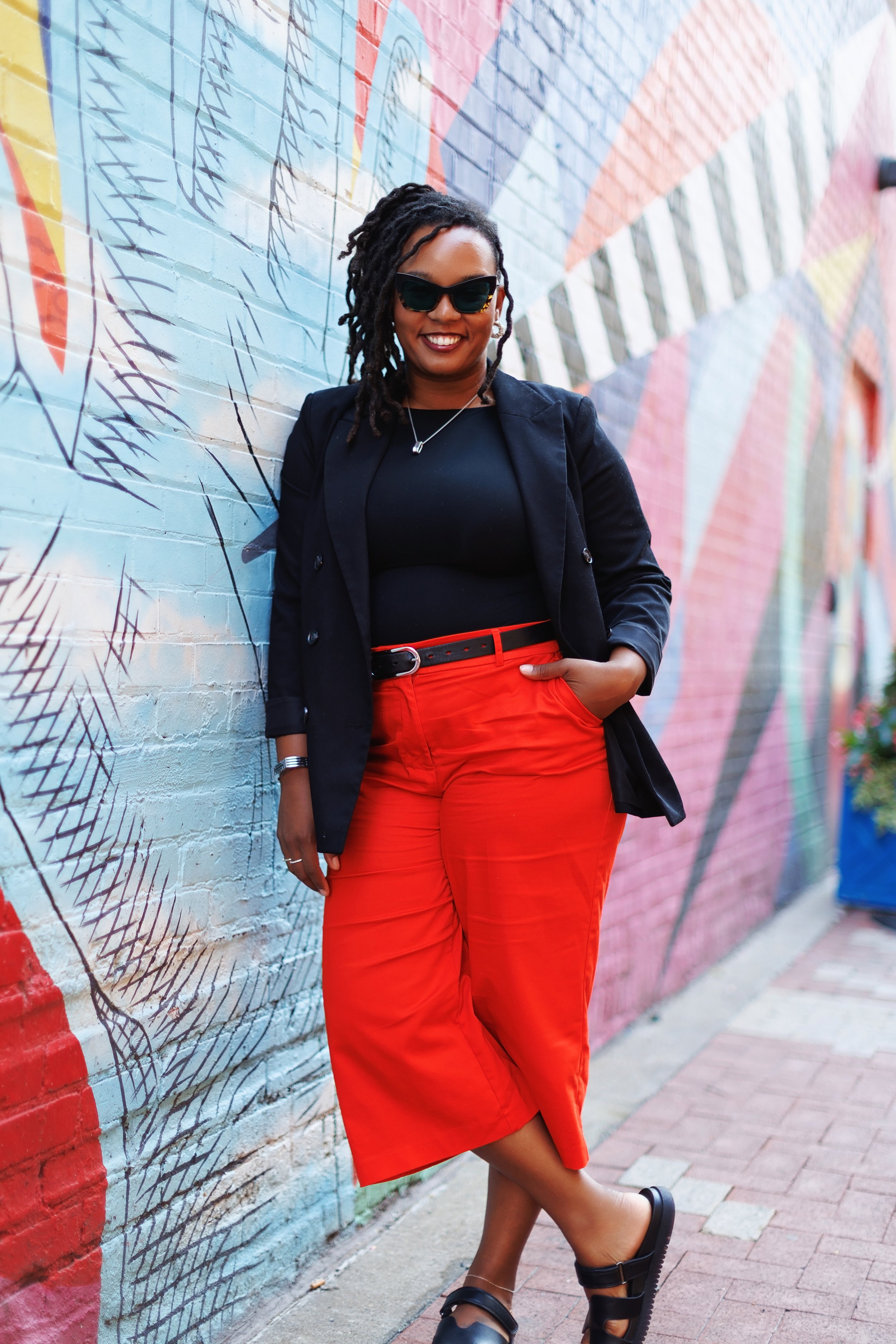 A woman with dreadlocks wearing sunglasses, a black blazer, a black top, red pants, and black sandals, standing with her hands in her pockets against a colorful graffiti wall.