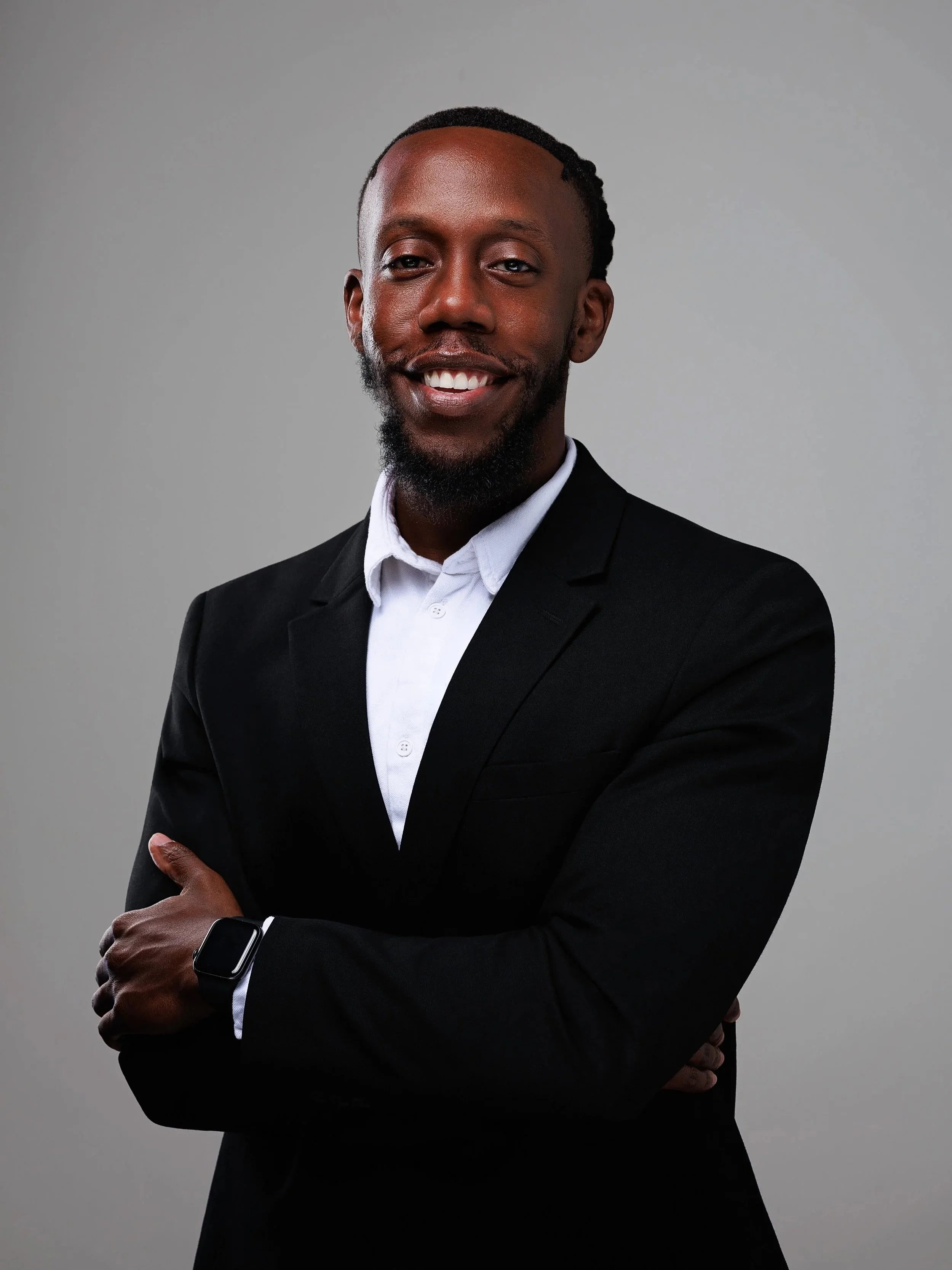 A smiling man in a black suit with a white shirt, standing against a gray background with arms crossed.