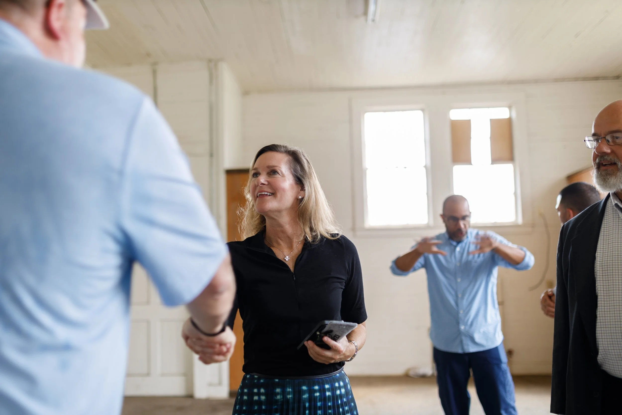 Group of people in a room, two people shaking hands, woman holding a phone, others engaging in conversation.