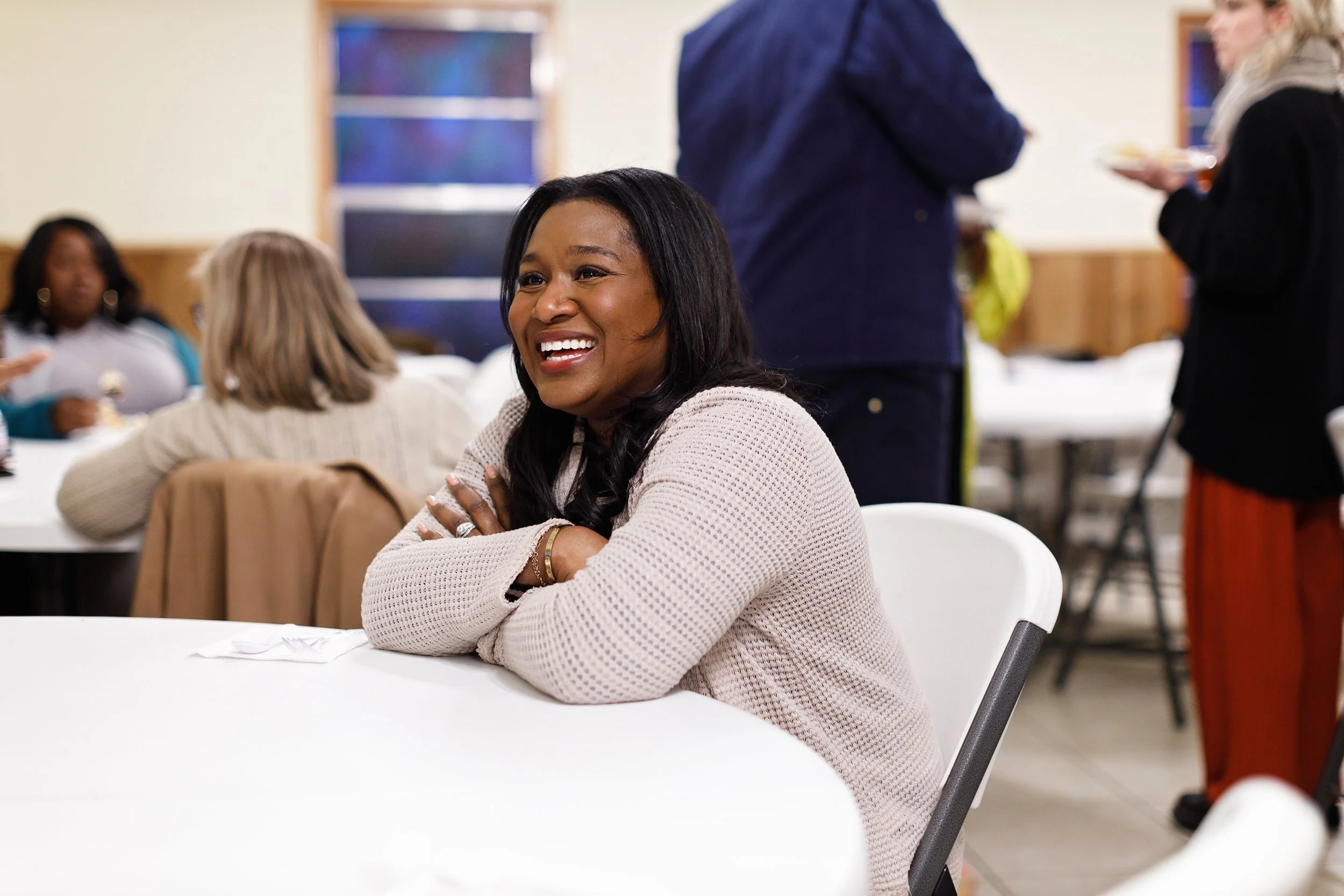 A woman smiling and sitting at a table during a social gathering or event, with other people in the background