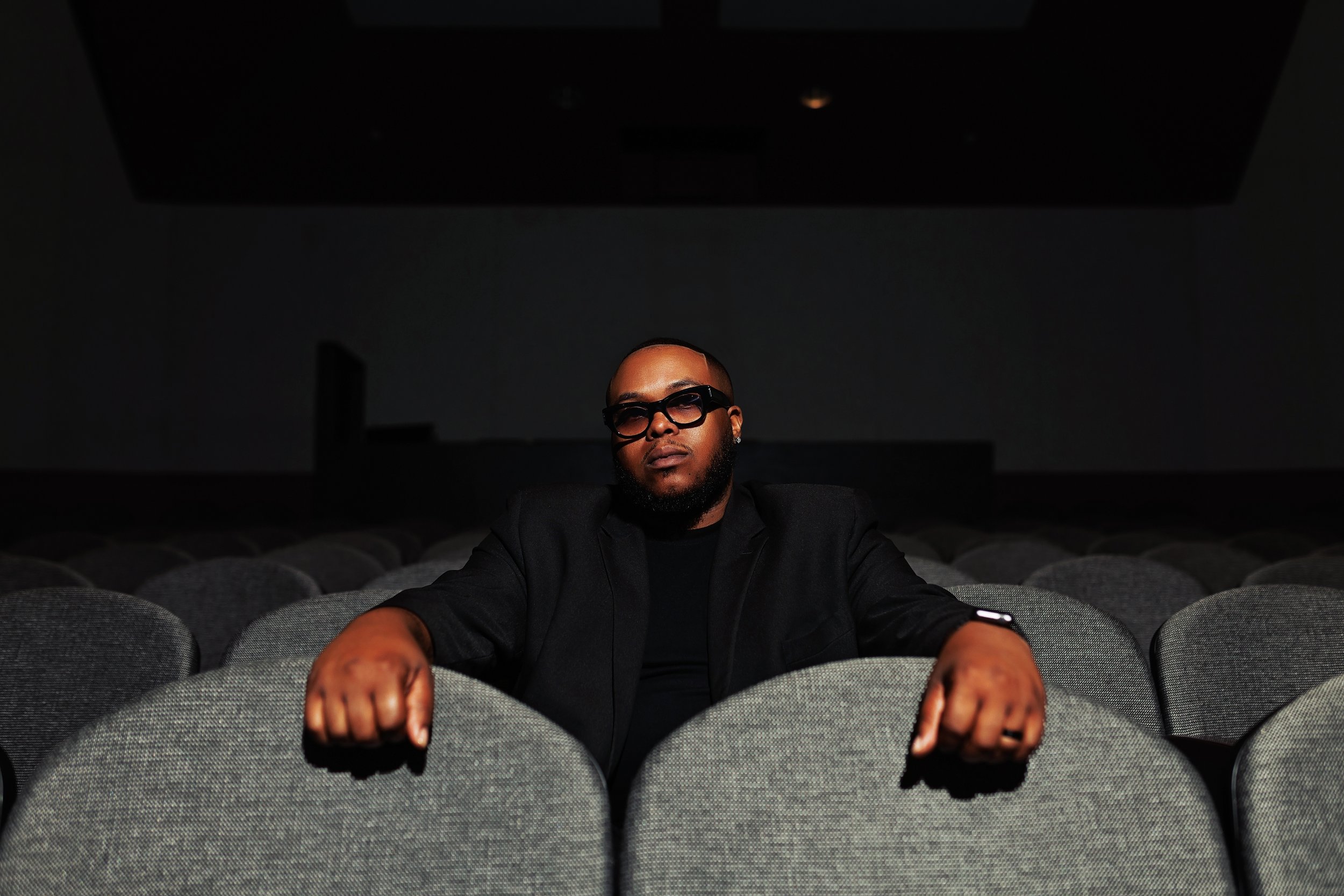A man wearing glasses, a dark shirt, and a black jacket sitting alone in an empty, dimly lit movie theater with rows of gray seats.