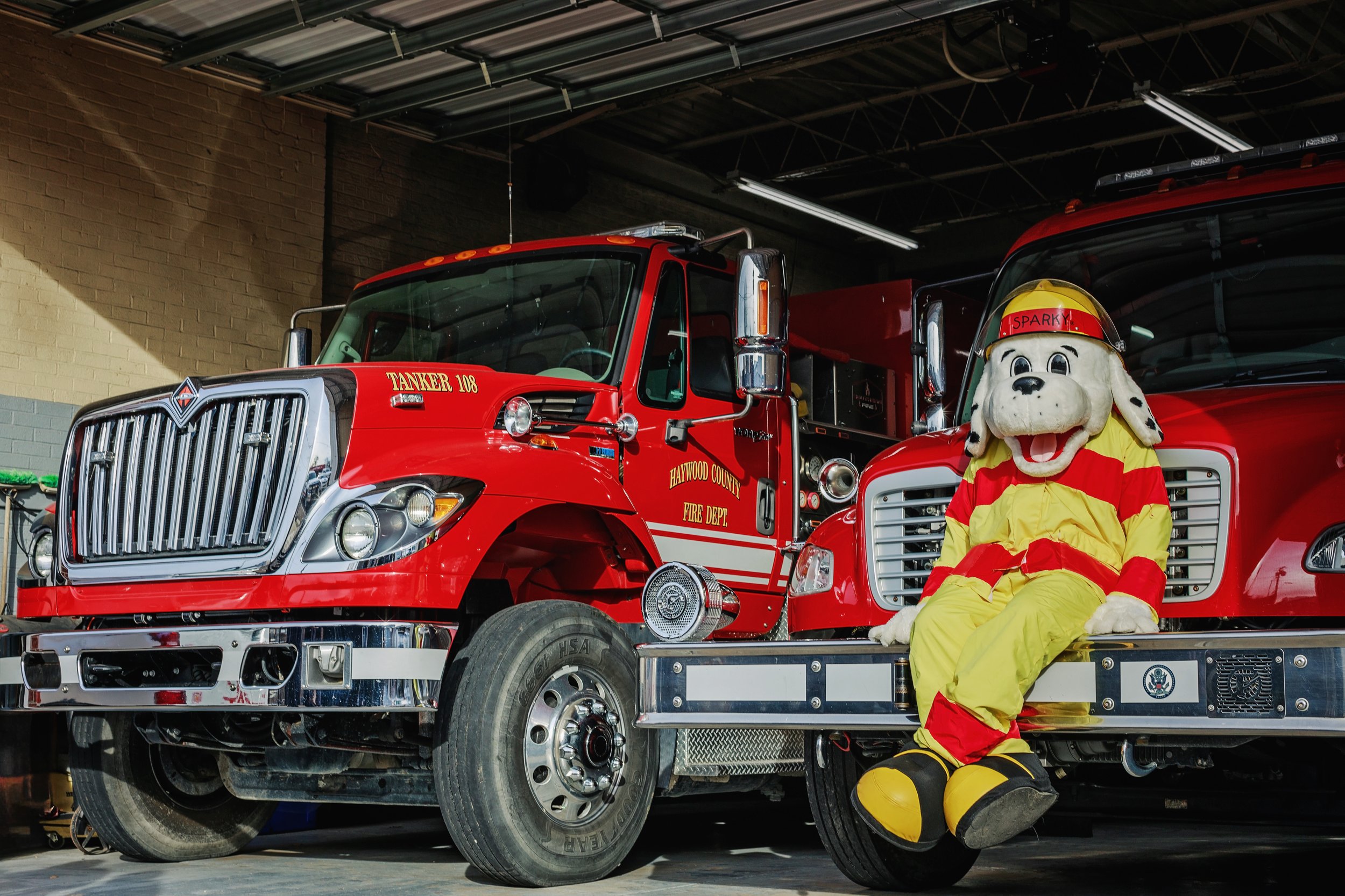 A fire truck from the Haywood County Fire Department parked inside a fire station with a mascot dressed as a dog in a firefighter uniform sitting on the bumper.