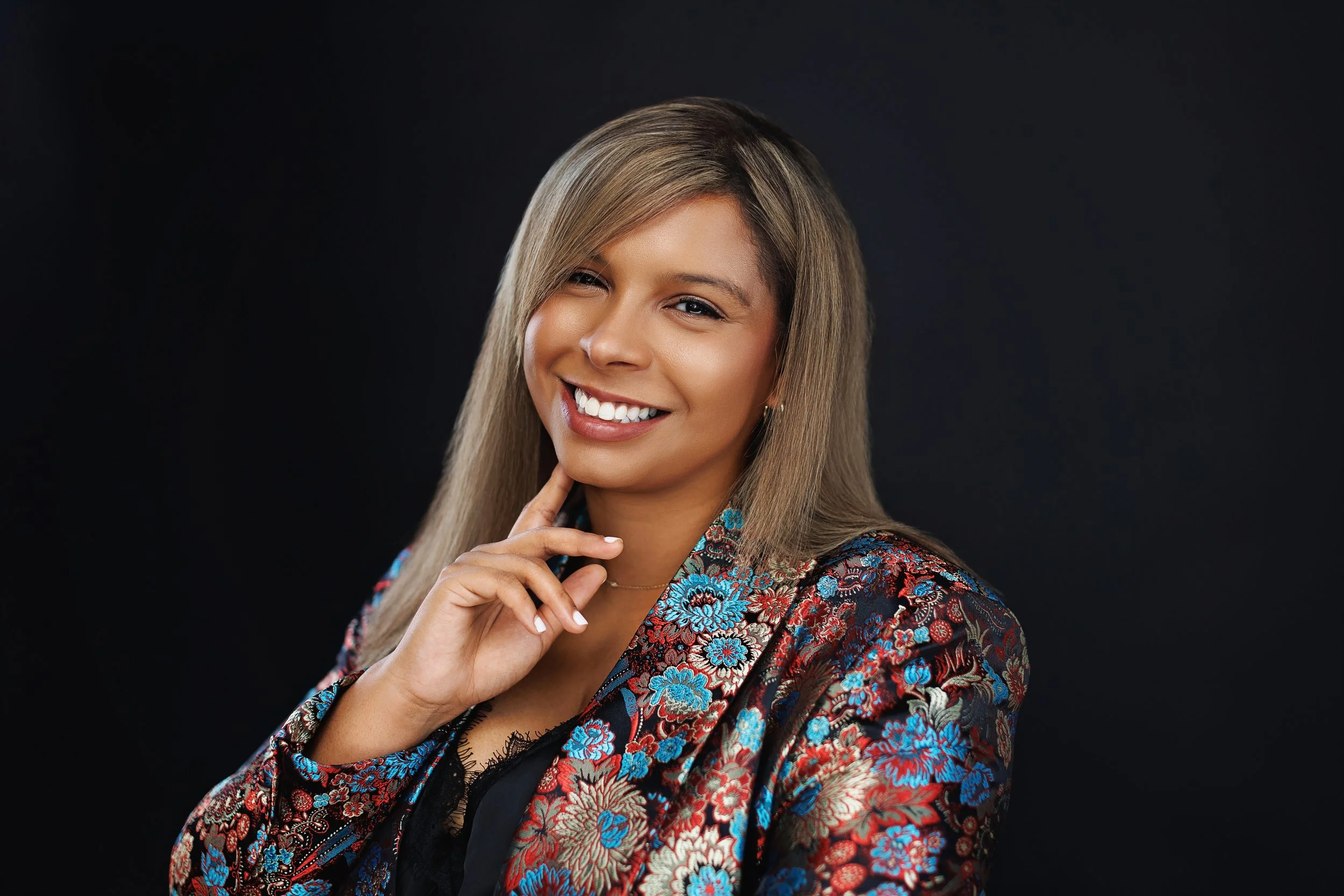 A smiling woman with light brown hair, wearing a black top and a colorful floral jacket, posing against a dark background.